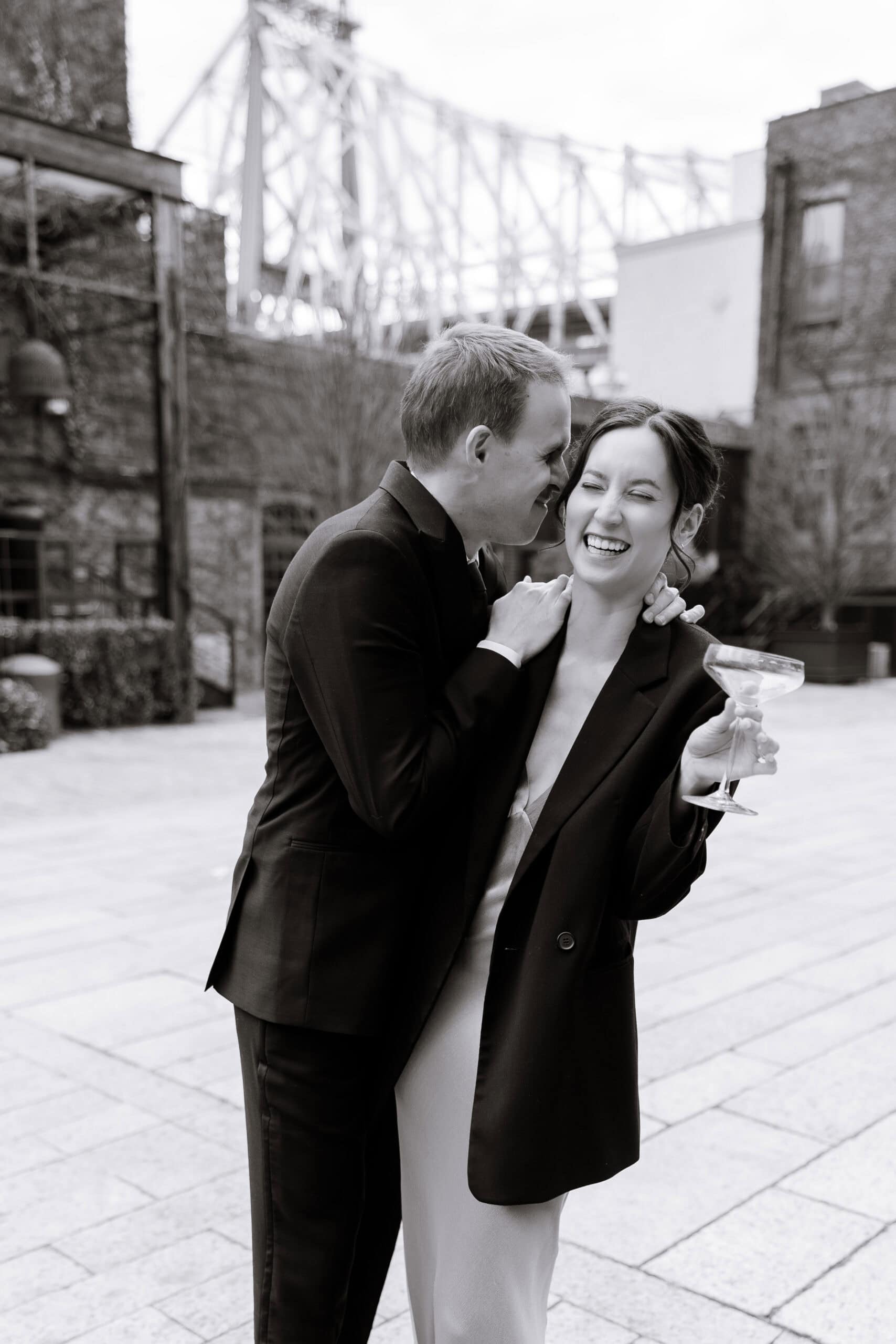 Black-and-white portrait of a couple embracing in an urban courtyard at The Foundry, with a bridge in the background and a coupe cocktail glass in hand.