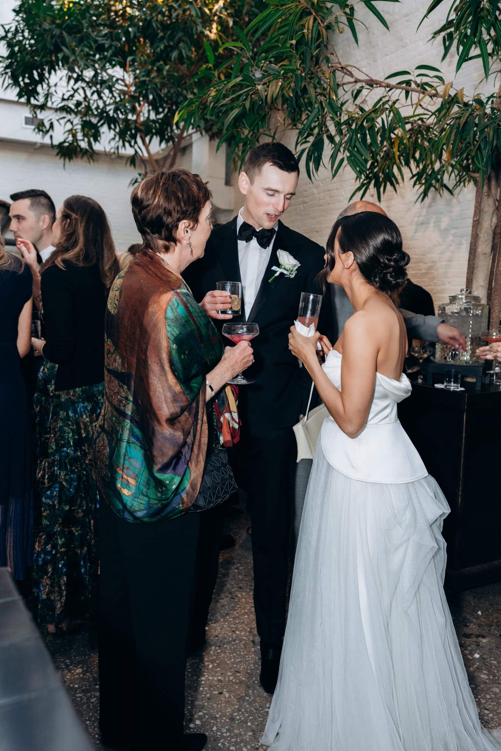 Bride in a strapless white gown and groom in a black tuxedo speaking with a guest during cocktail hour indoors at The Foundry, surrounded by greenery and drinks.