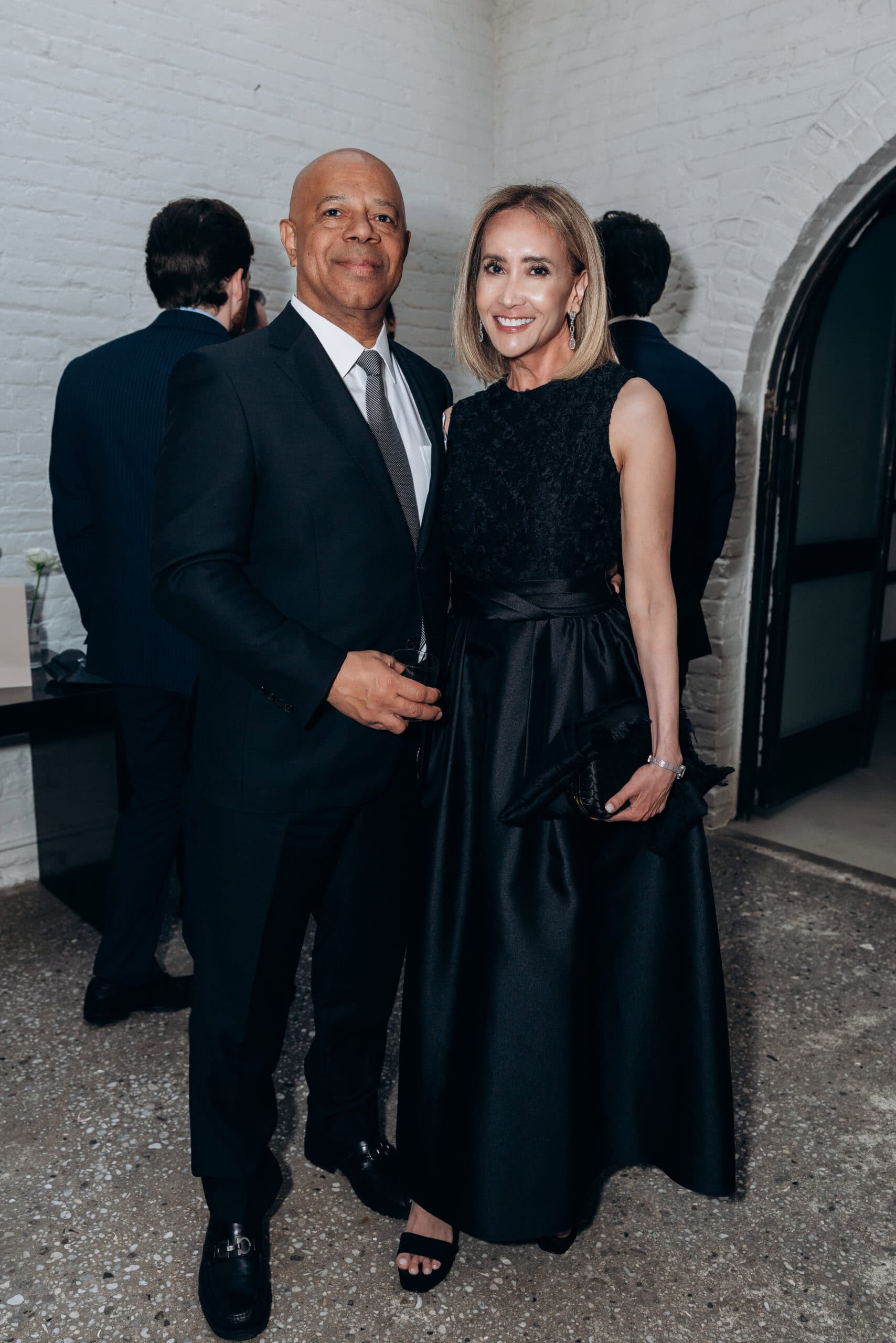 Two formally dressed wedding guests pose indoors at The Foundry, standing before white brick walls and an arched doorway during a black-tie reception.