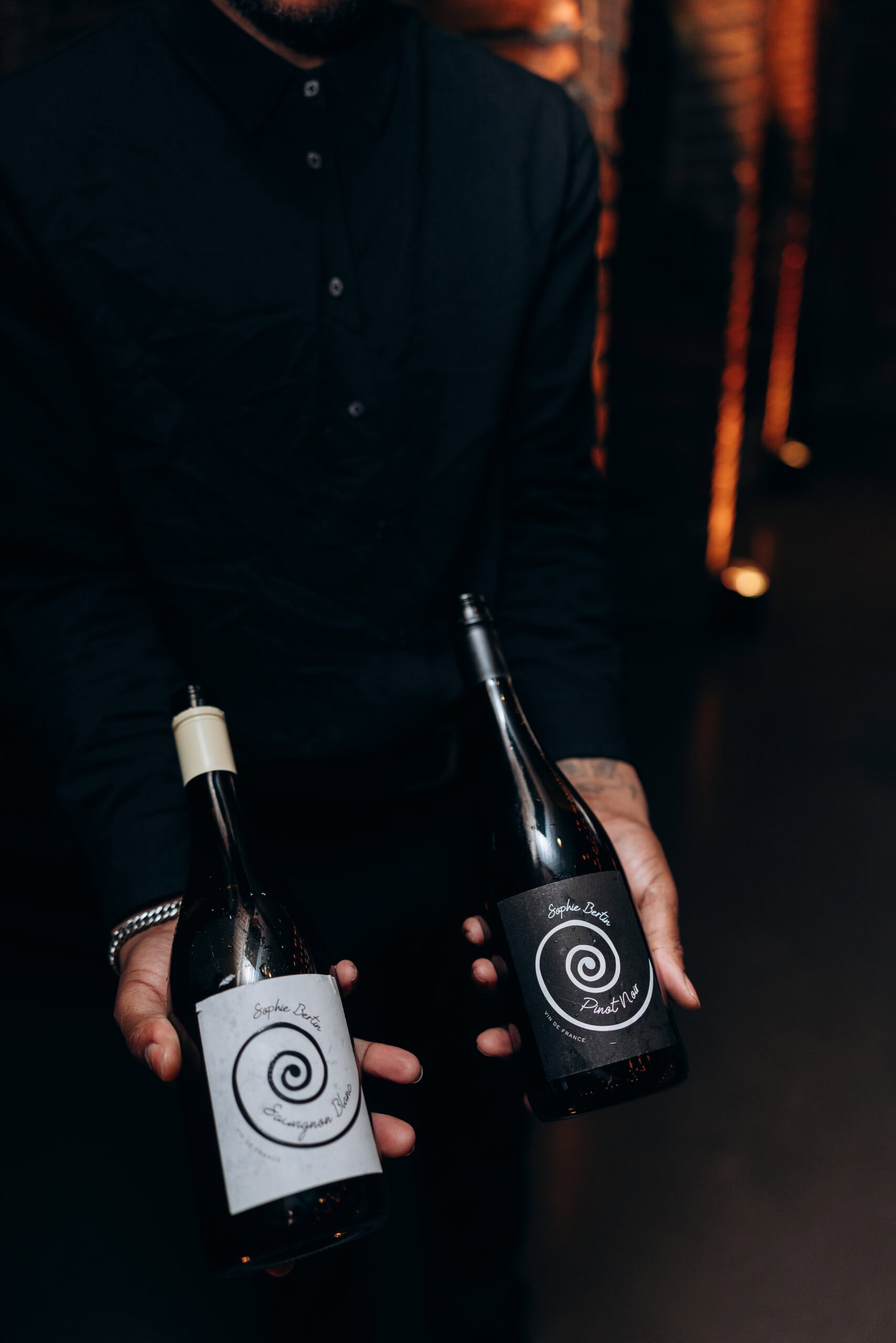 At The Foundry, a person in a black shirt holds two Sophie Bertin wine bottles—Sauvignon Blanc and Pinot Noir—in a dimly lit interior.