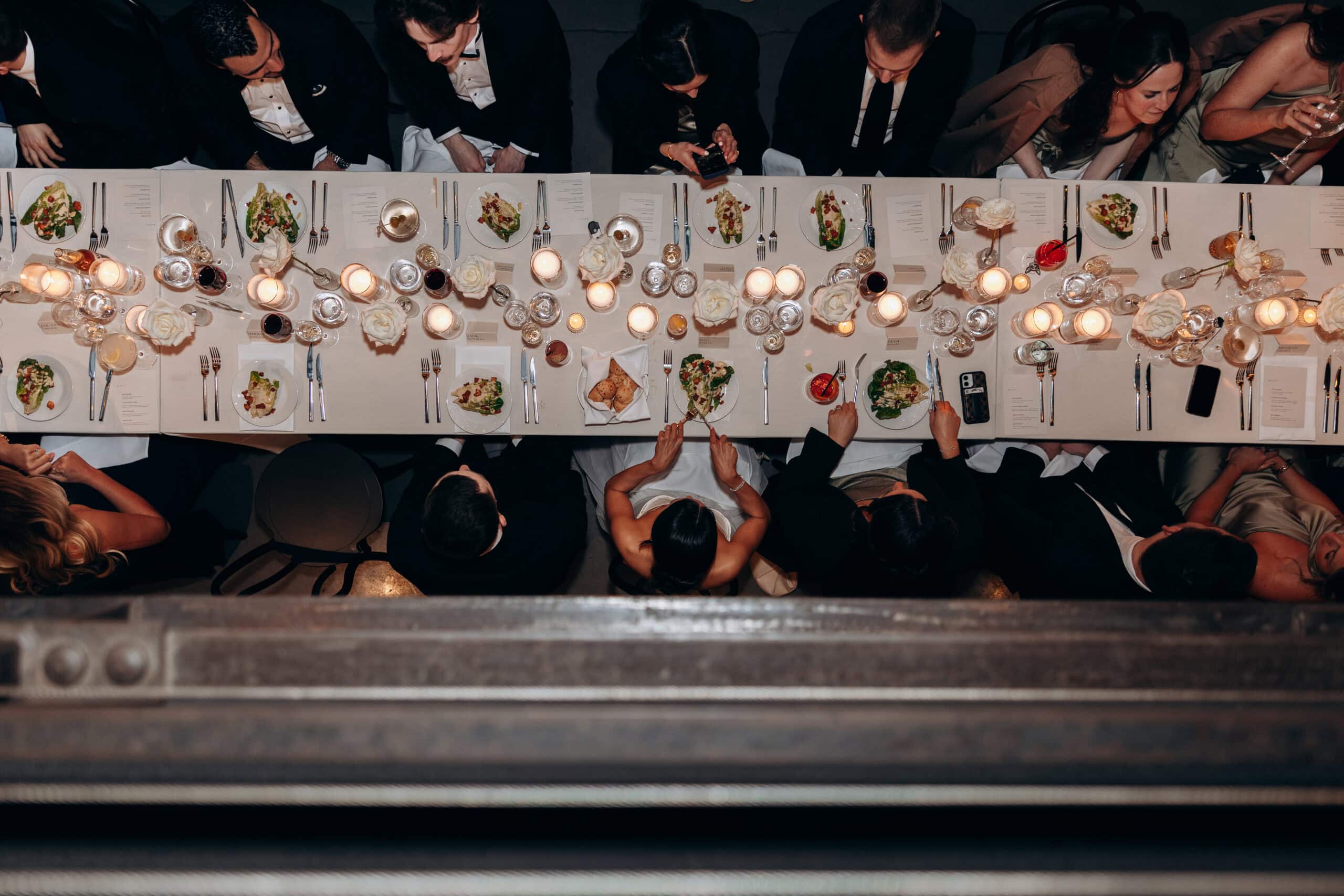 Overhead view of guests seated at a long candlelit reception table at The Foundry, set with white roses, plated salads, menus, glassware, and black-tie attire.