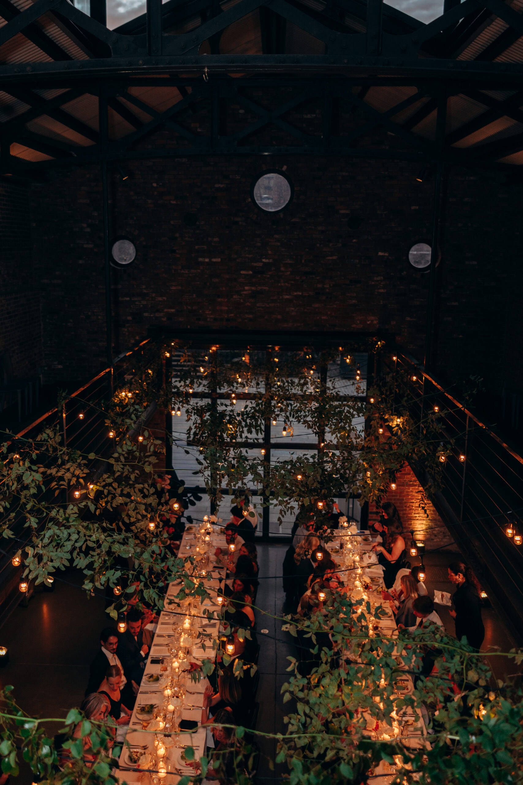 Overhead view of a wedding reception dinner at The Foundry, with long candlelit tables, hanging greenery, string lights, exposed brick, and dark steel rafters.