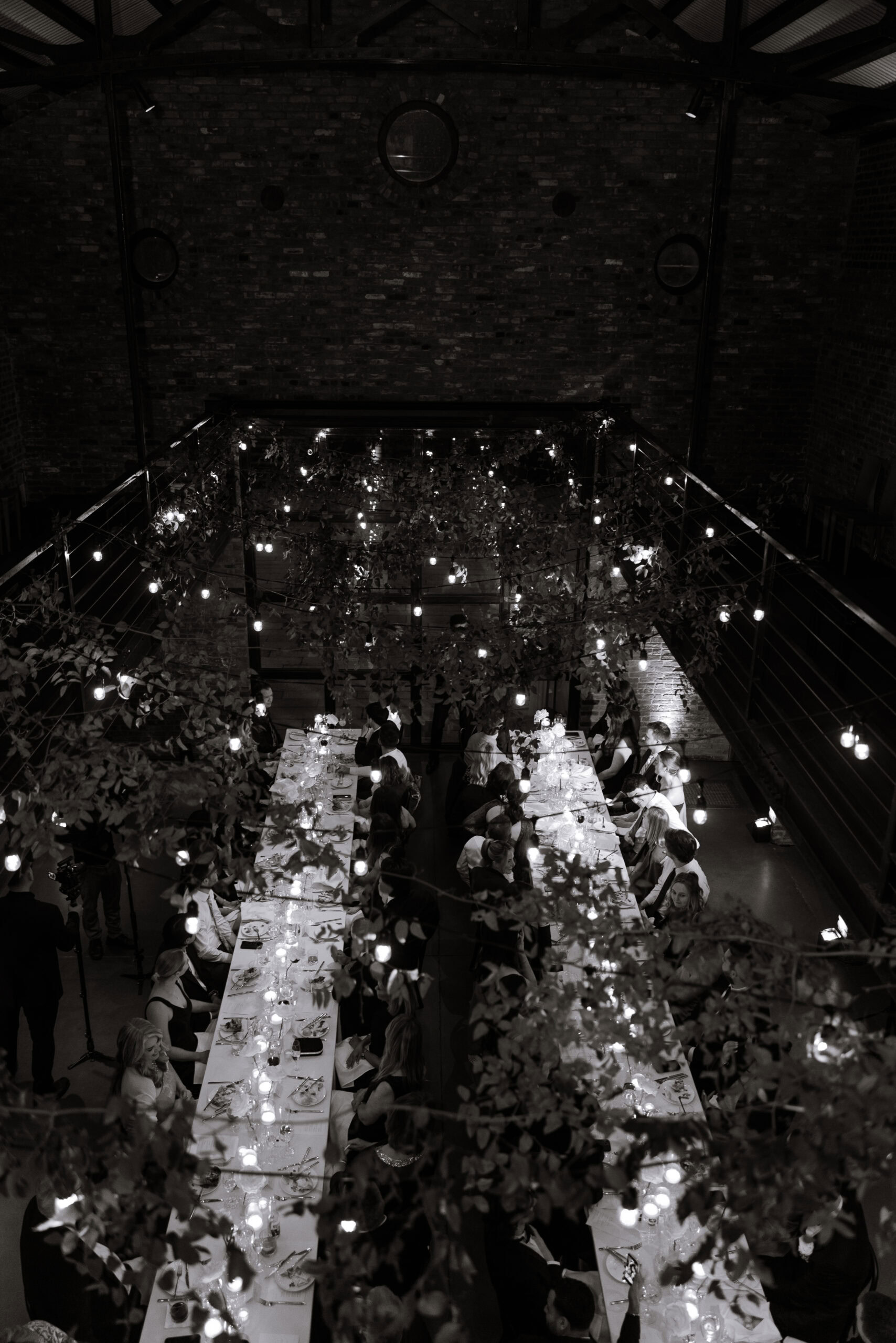 Black-and-white overhead view of a wedding reception dinner at The Foundry, with long candlelit tables, hanging greenery, string lights, and guests in an industrial brick space.