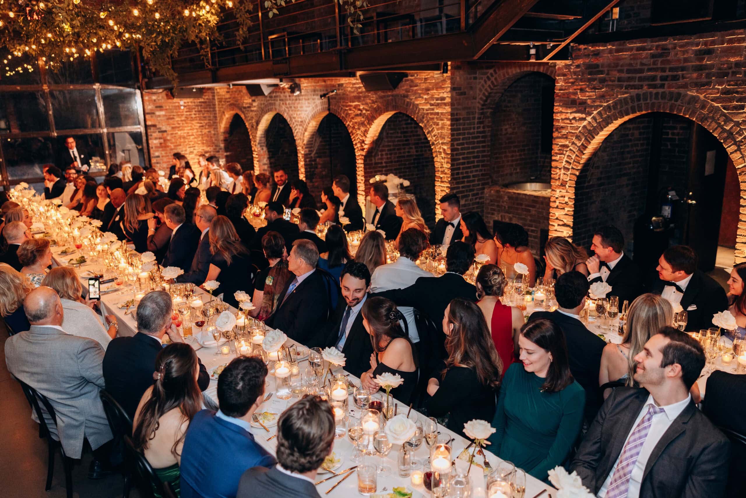 Wedding guests seated at long candlelit tables during a reception at The Foundry, surrounded by exposed brick arches, string lights, and white floral arrangements.