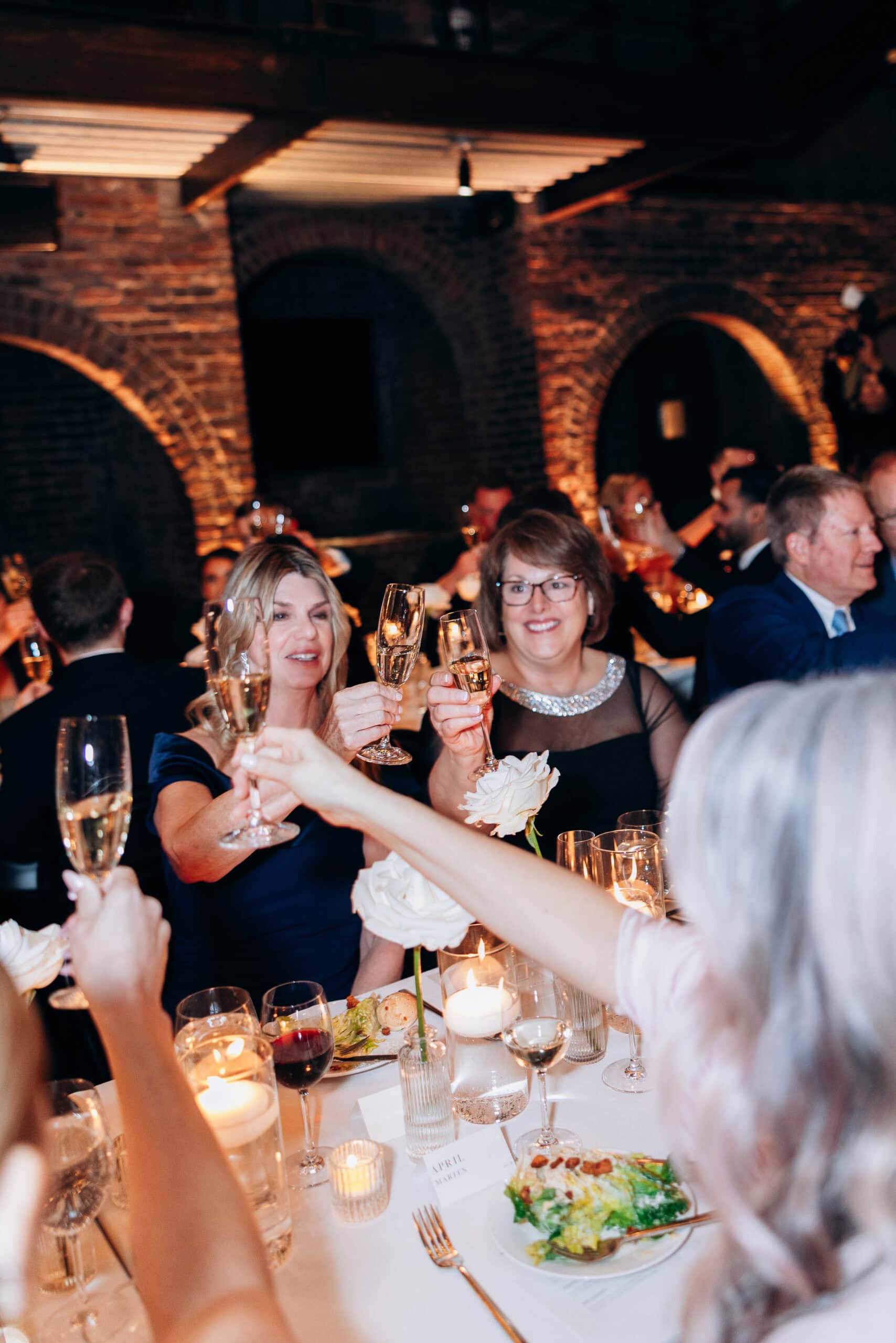 Wedding guests toast with champagne at a candlelit reception table inside The Foundry, framed by exposed brick arches, white roses, and glowing votives.