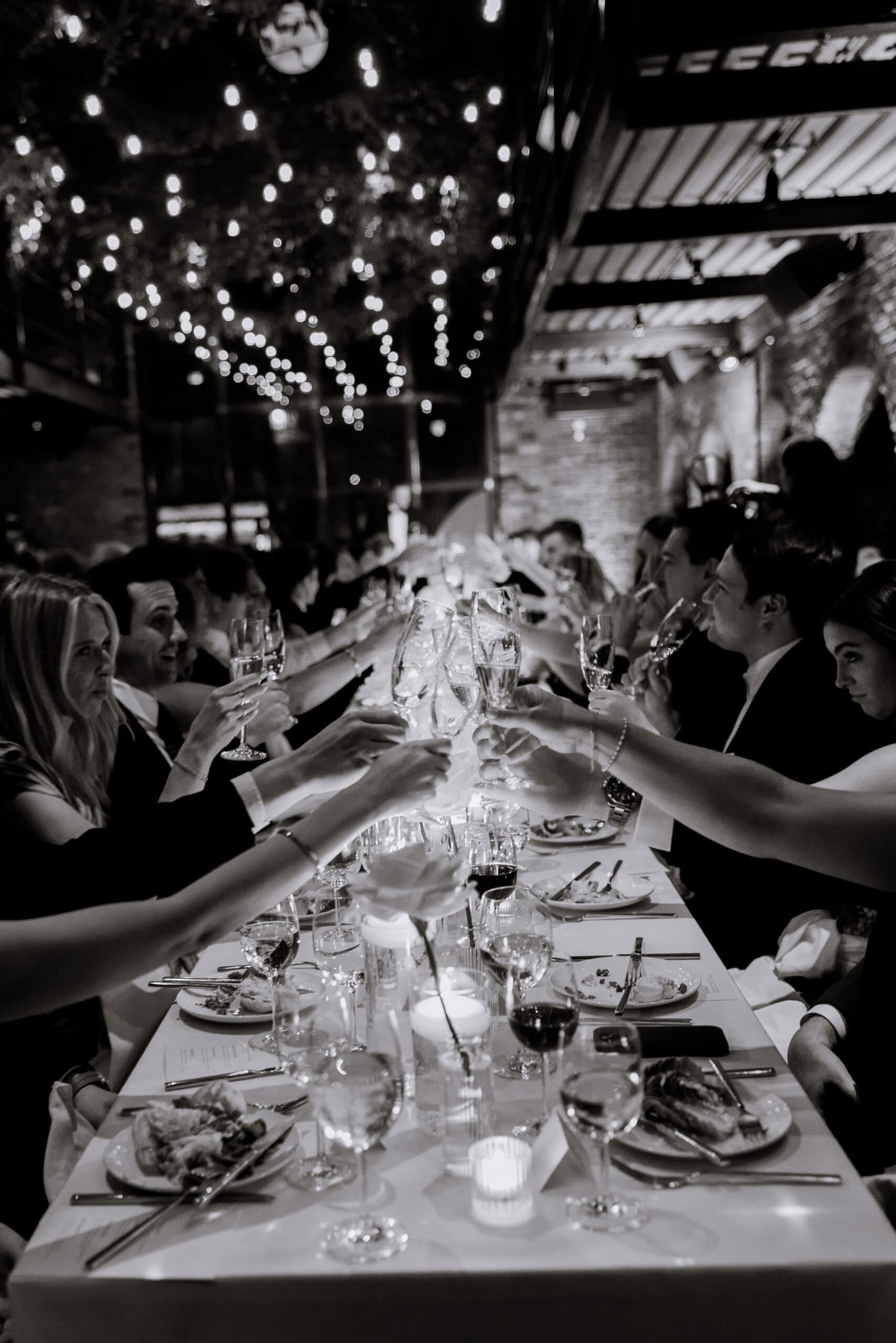 Black-and-white wedding reception at The Foundry with guests raising champagne glasses over a candlelit long table beneath string lights in an industrial brick space.