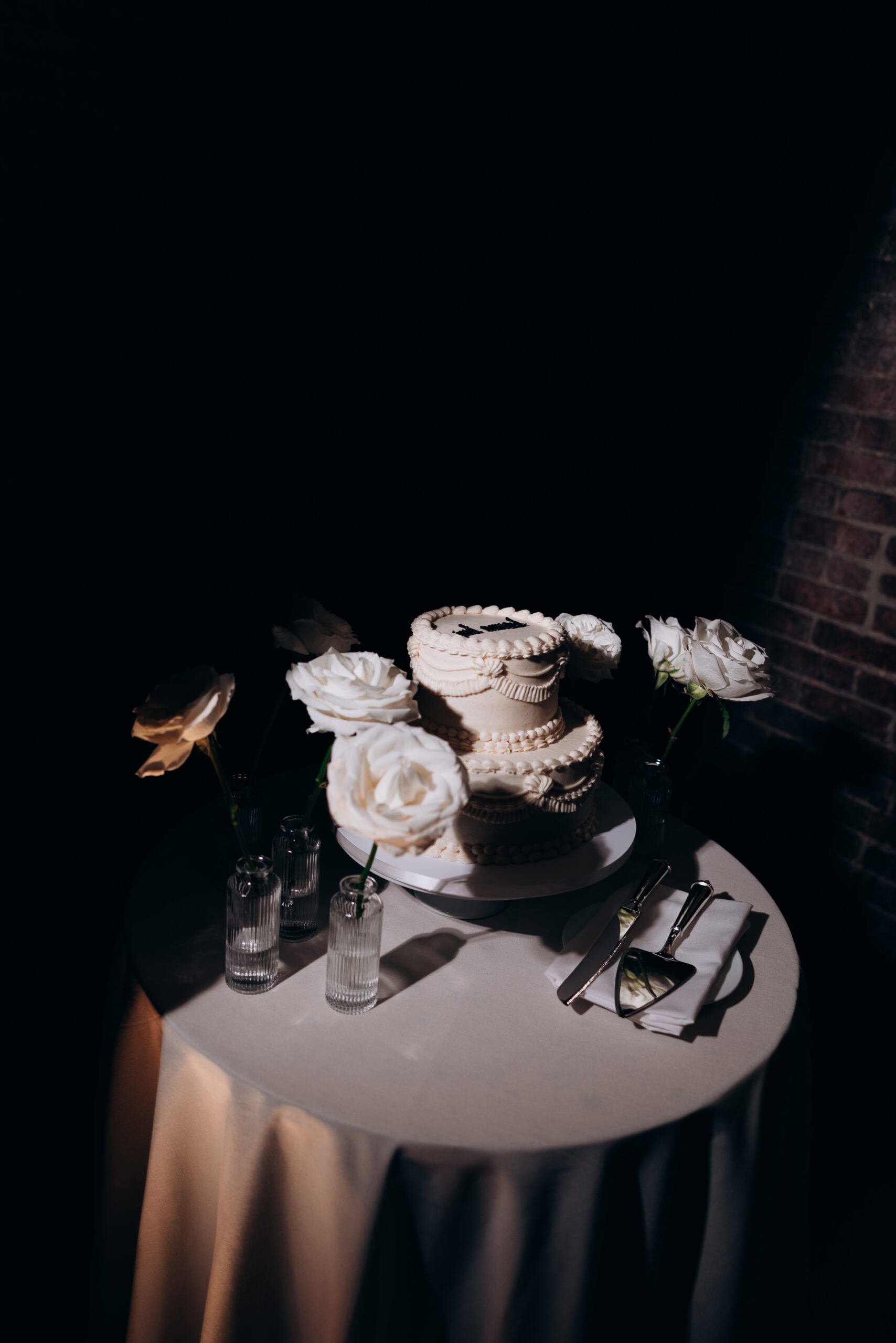 Spotlit white tiered wedding cake on a round table with white roses in glass bud vases, cake knife and server, and a dark brick backdrop at The Foundry.