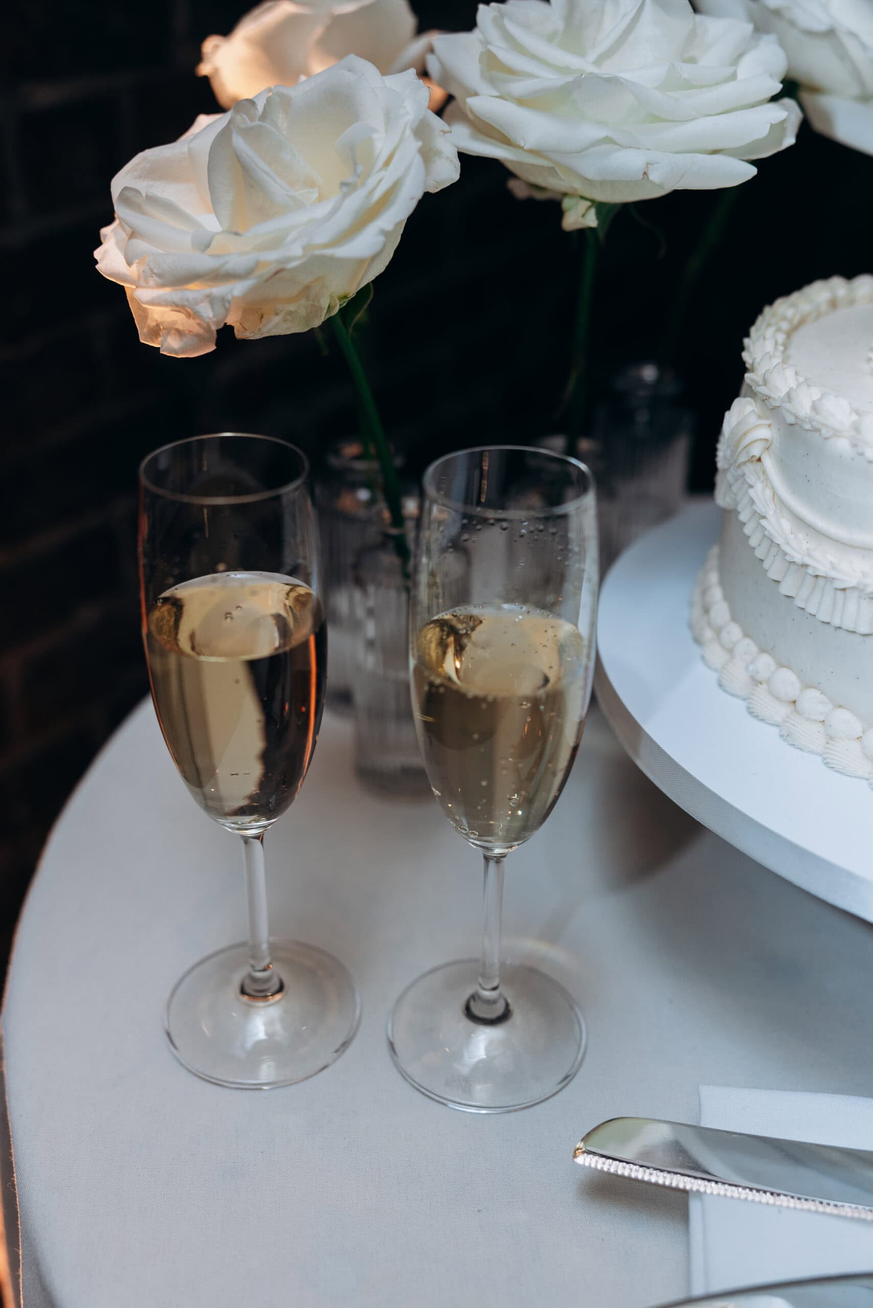 Close-up of a wedding reception table at The Foundry with two champagne flutes, white rose stems in glass vases, and a white frosted cake.