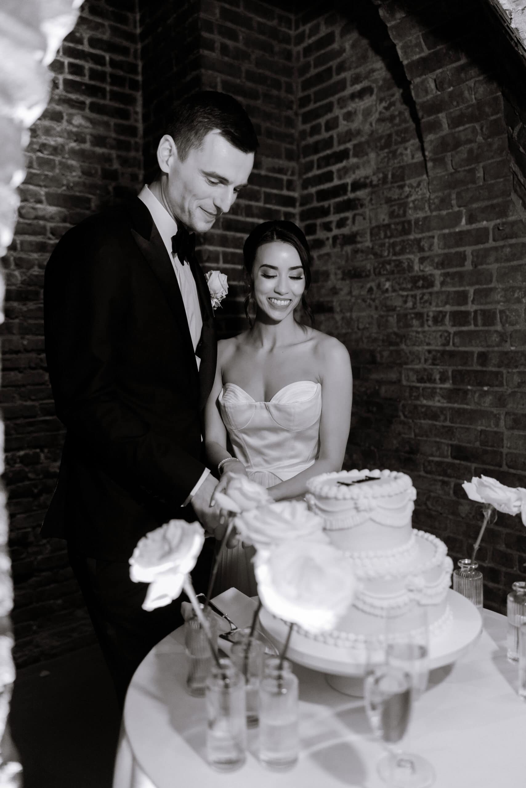 Black-and-white wedding reception portrait of a couple cutting a tiered cake beside white roses under brick arches at The Foundry.