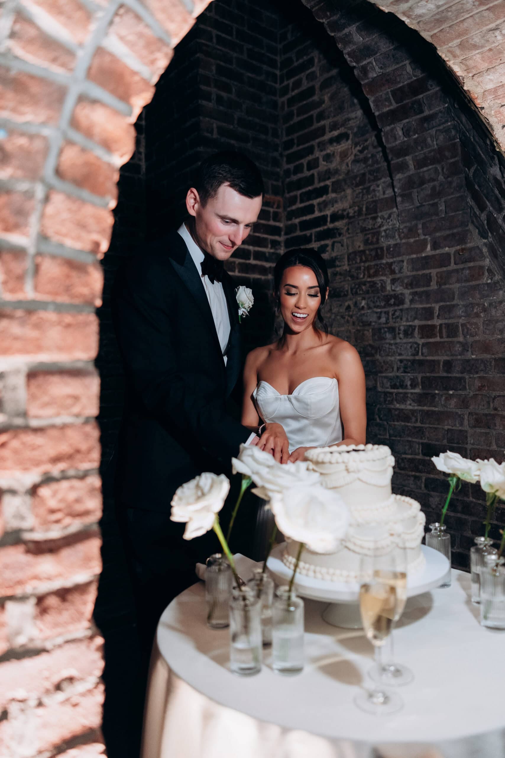 Bride and groom cutting a tiered white wedding cake under brick arches at The Foundry, with white roses in bud vases and champagne on a round table.
