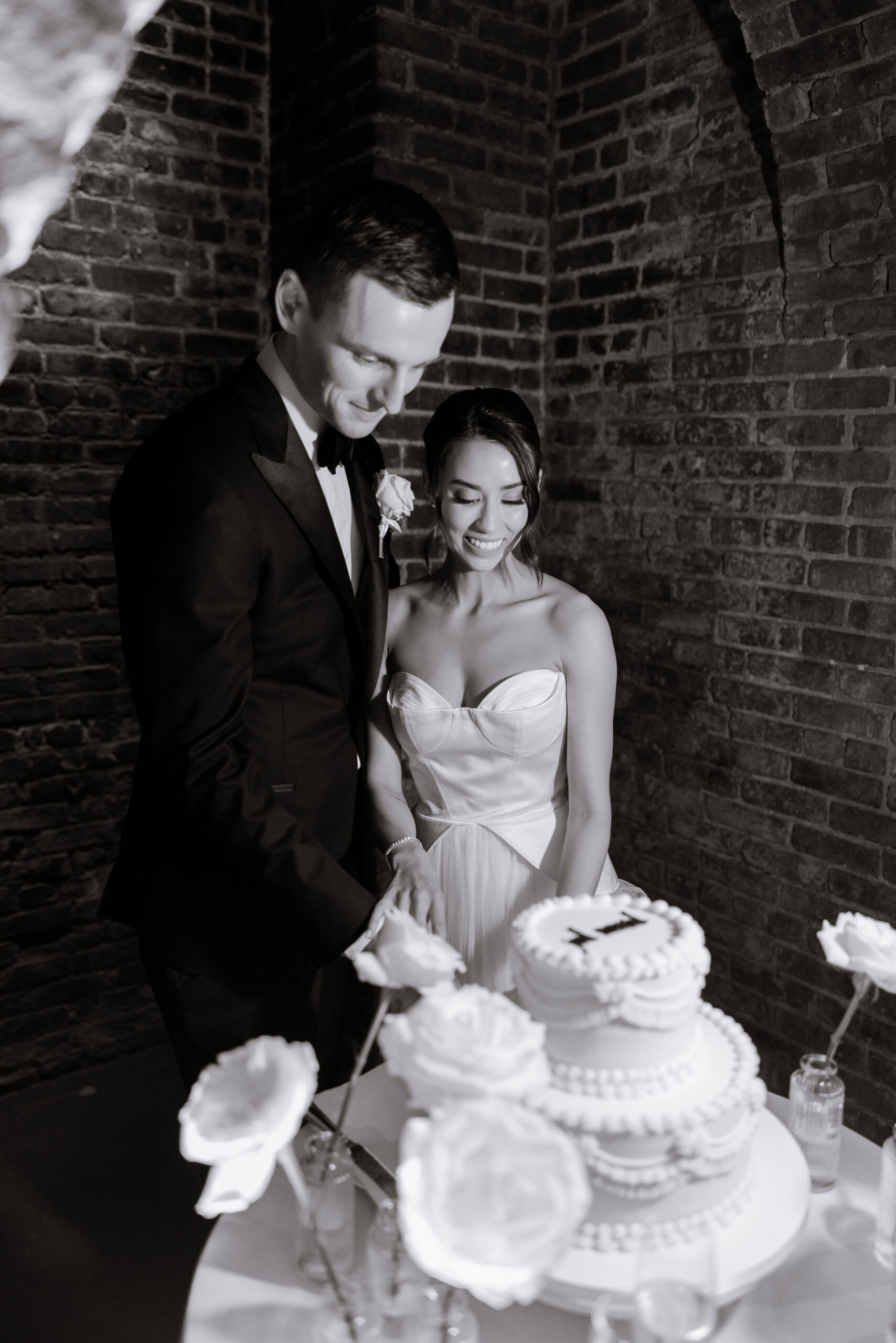 Black-and-white wedding portrait of a couple cutting a tiered cake at The Foundry, surrounded by brick arches, white florals, and soft reception lighting.