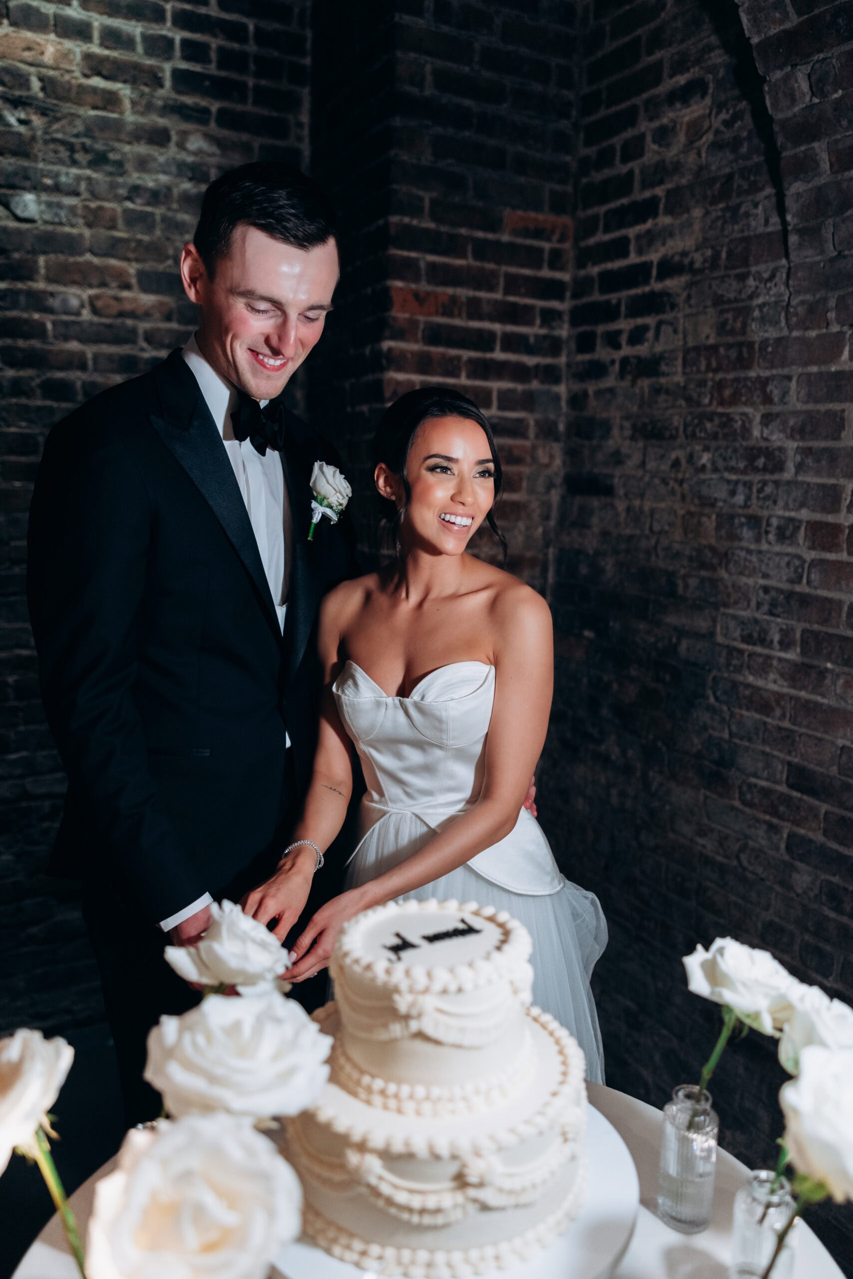 Bride and groom cut a tiered white wedding cake surrounded by white roses against exposed brick walls at The Foundry.