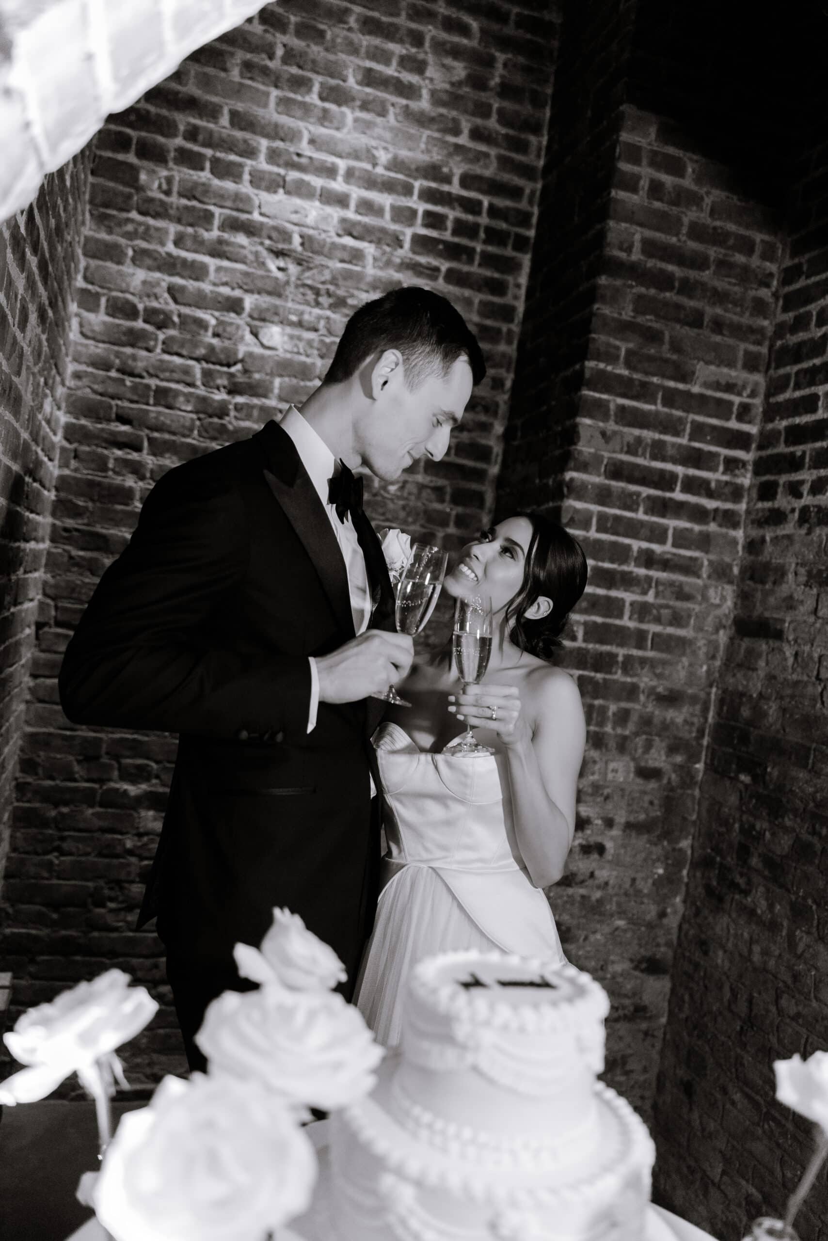 Bride and groom toast with champagne beside a tiered cake under brick arches at The Foundry in a black-and-white wedding portrait.