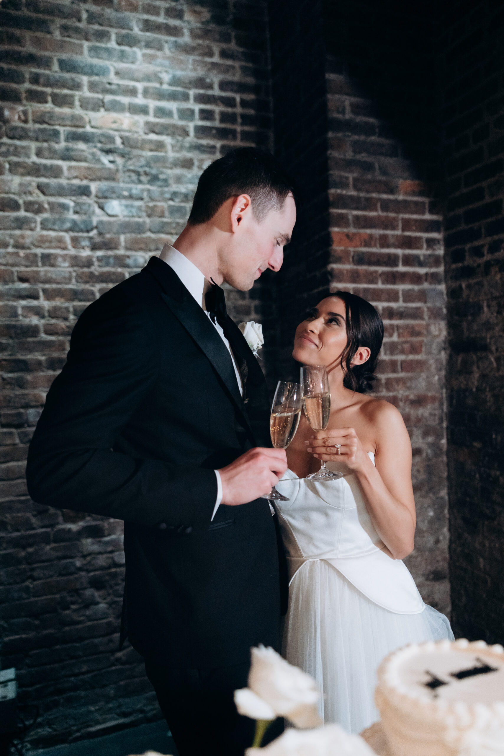 Bride and groom at The Foundry share a champagne toast beside a cake, standing against exposed brick walls in evening light.