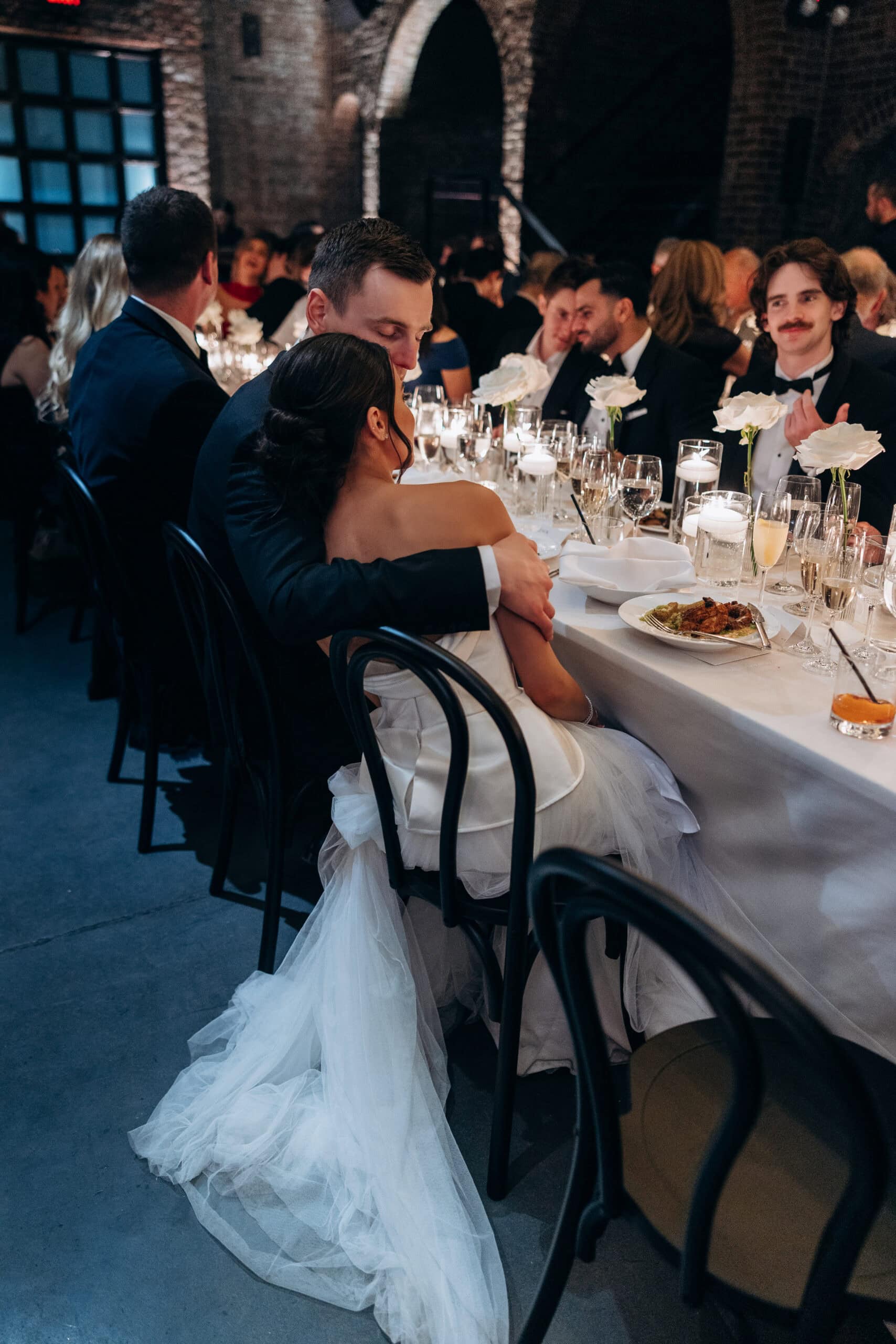 Bride and groom seated together at a candlelit reception table at The Foundry, surrounded by guests, white roses, glassware, and dramatic brick arches.
