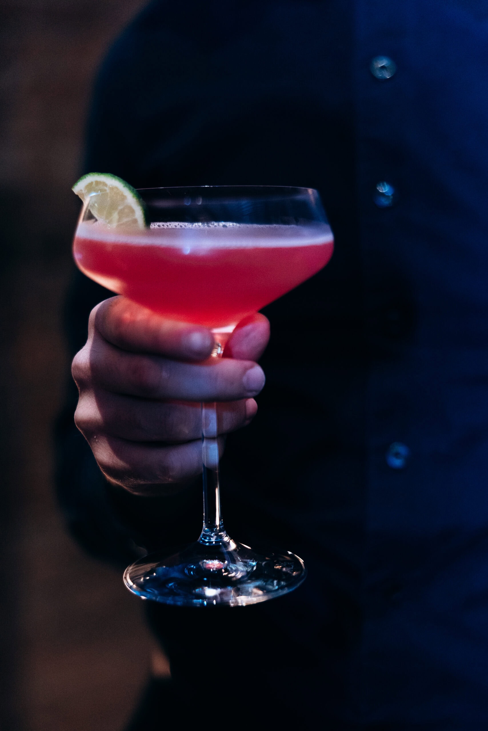 Close-up of a guest in a dark suit holding a pink cocktail in a coupe glass with a lime garnish during wedding cocktail hour at The Foundry.