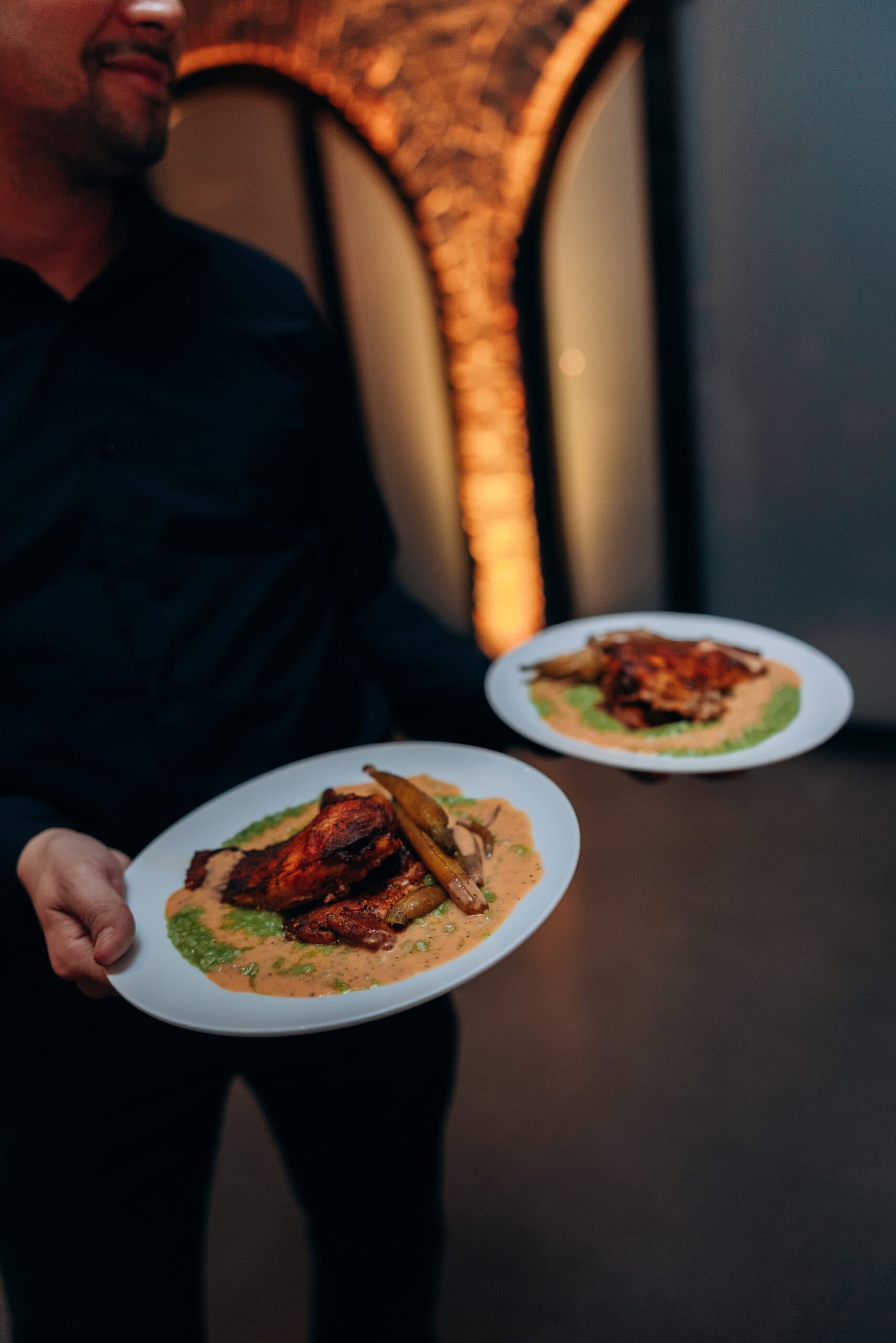 Server carrying two plated entrées with roasted poultry and vegetables in a warmly lit reception space at The Foundry, framed by glowing brick arches.