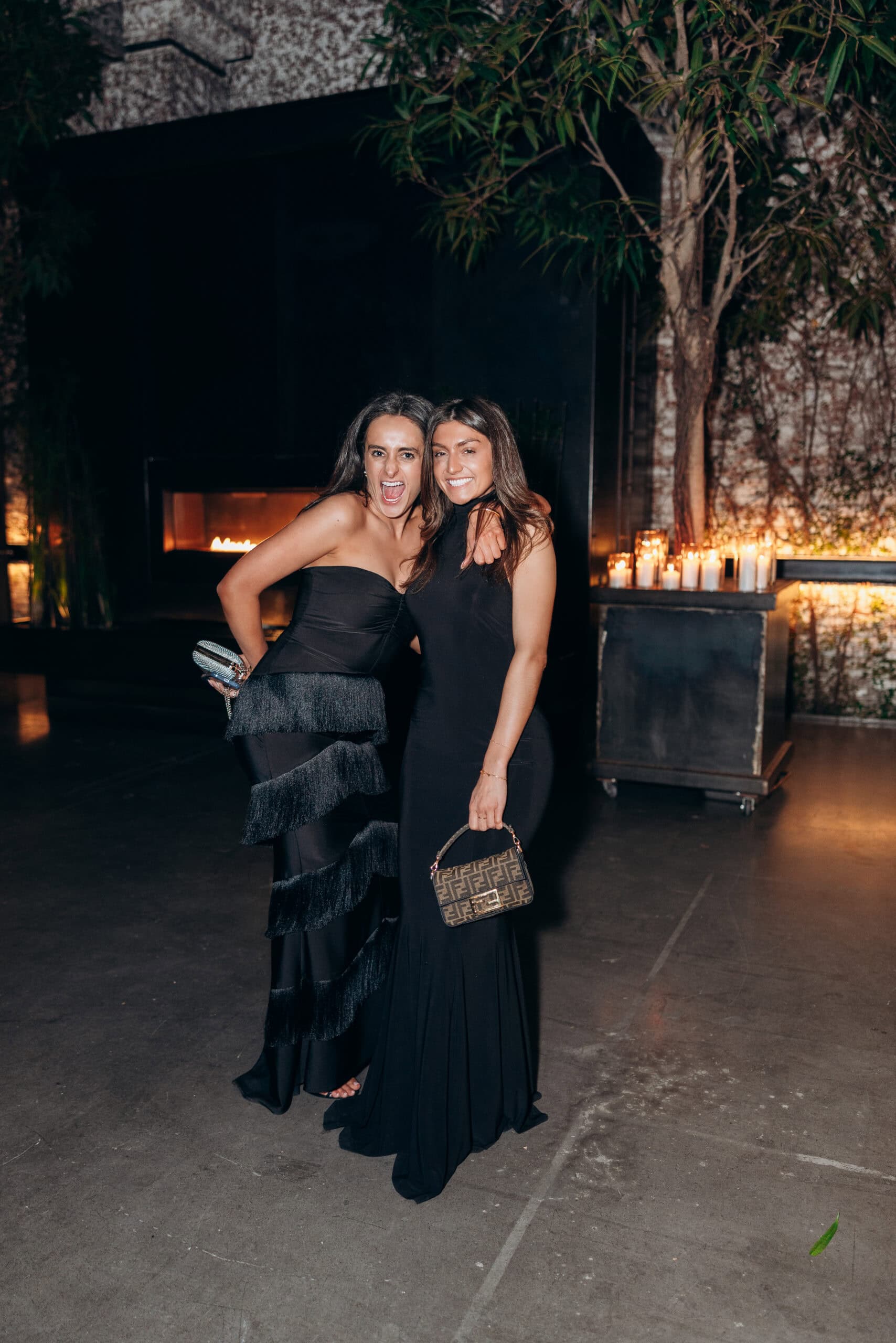 Two women in black formal gowns pose arm in arm at The Foundry, standing before a dark fireplace with candles and tall greenery at an evening wedding reception.