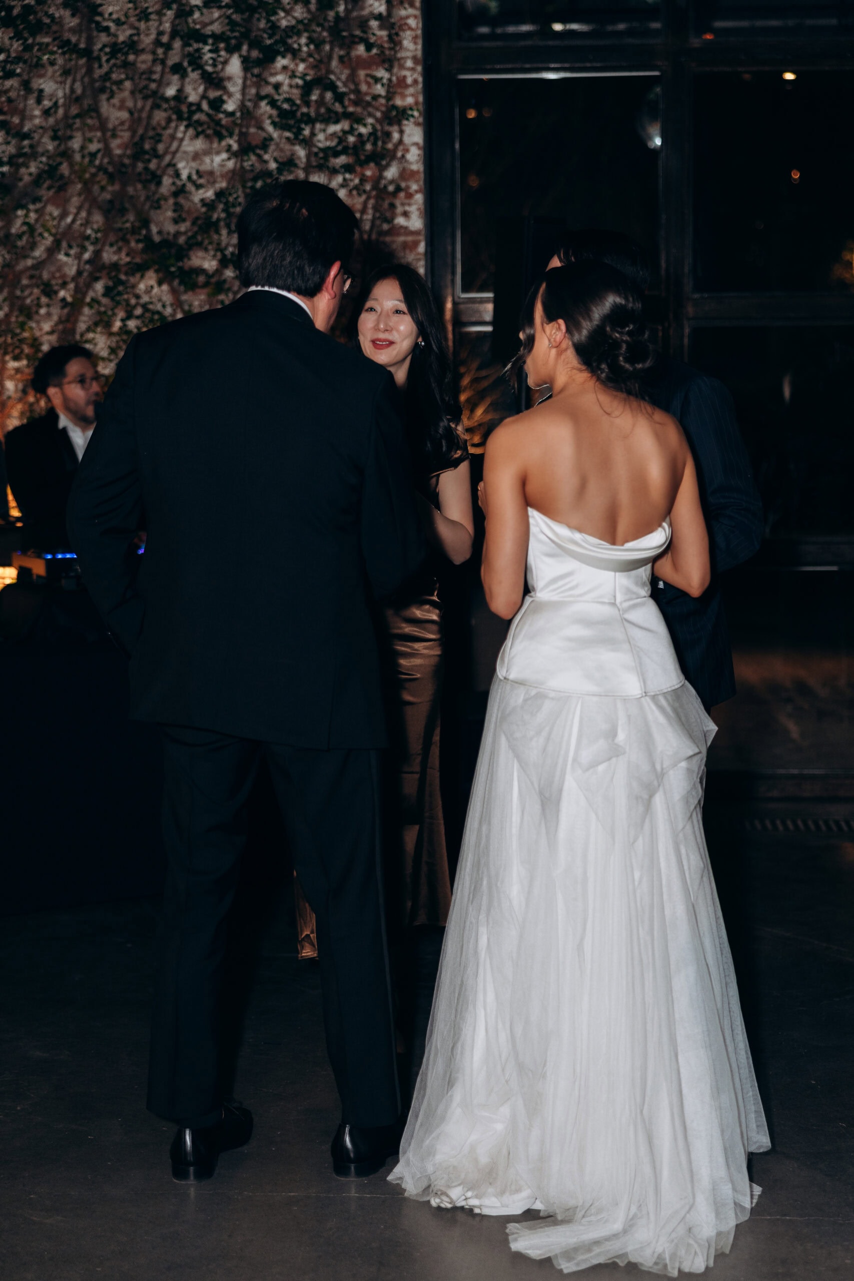 At The Foundry, a bride in a strapless white gown stands with guests in dark formalwear during an indoor evening wedding reception beside an ivy-covered brick wall.