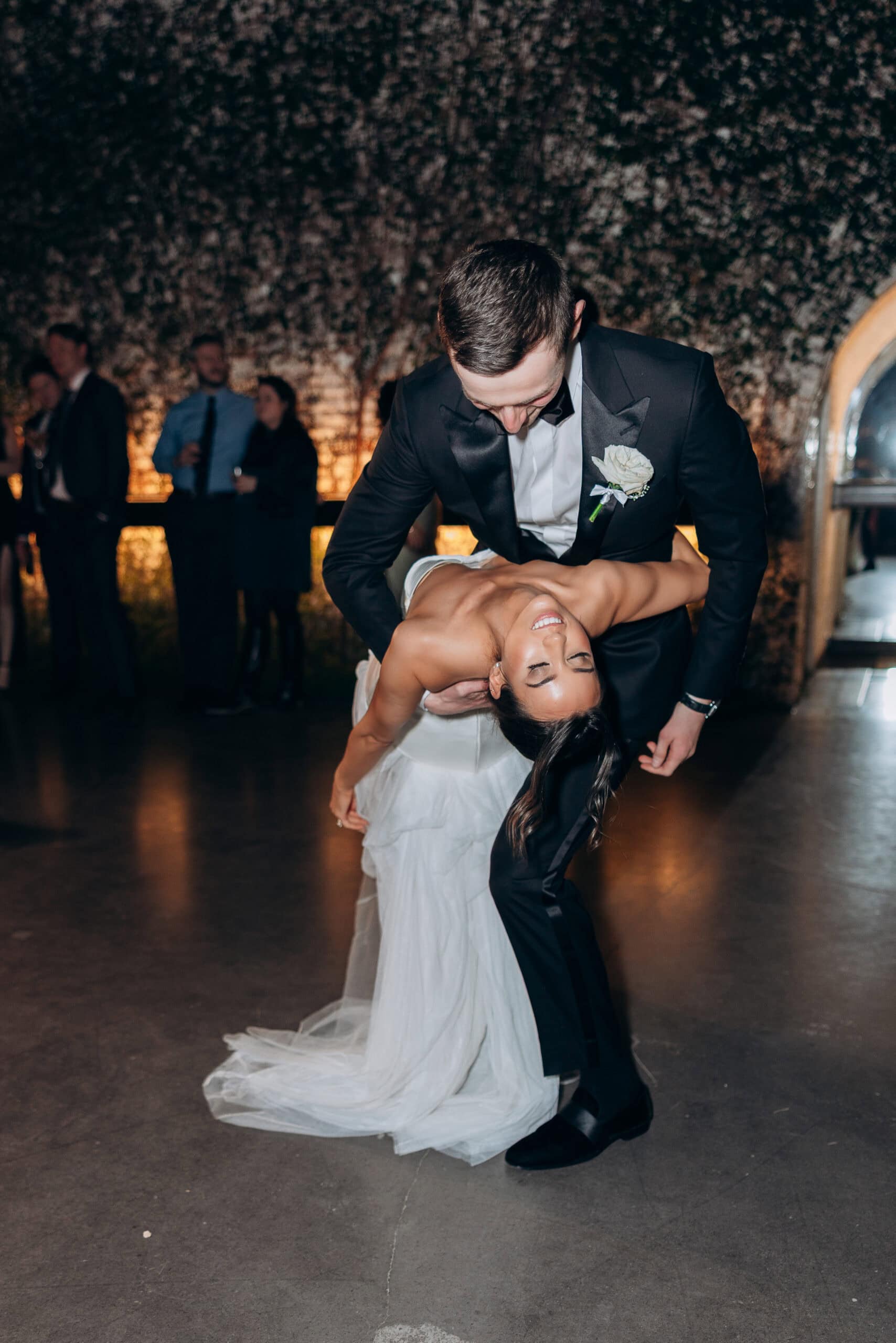 Bride and groom share a dramatic dip during their evening reception dance at The Foundry, with guests softly blurred behind them and an ivy-covered wall in warm light.