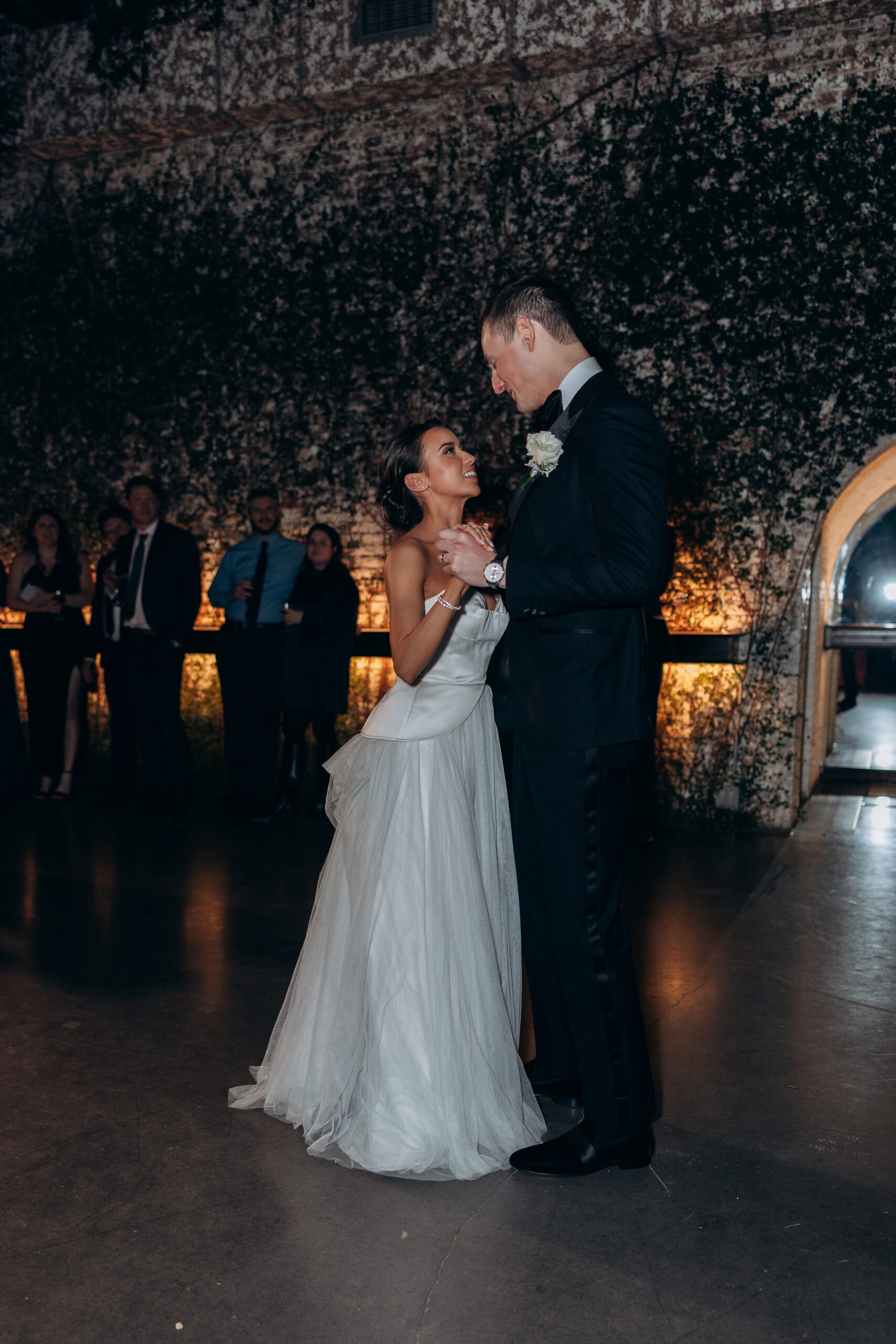 Bride and groom share their first dance at The Foundry, surrounded by ivy-covered brick walls, warm ambient light, and guests watching from the edges.