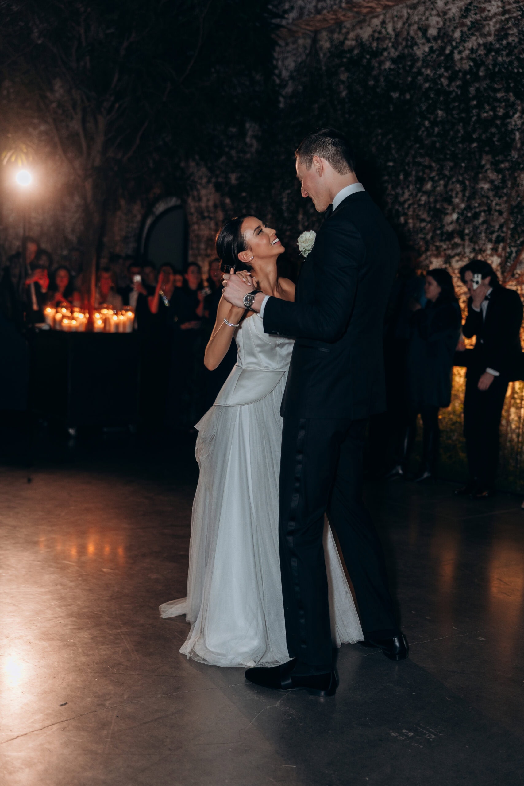 Bride and groom share their first dance at The Foundry in a dimly lit reception space with candlelight, stone walls, and guests watching in the background.