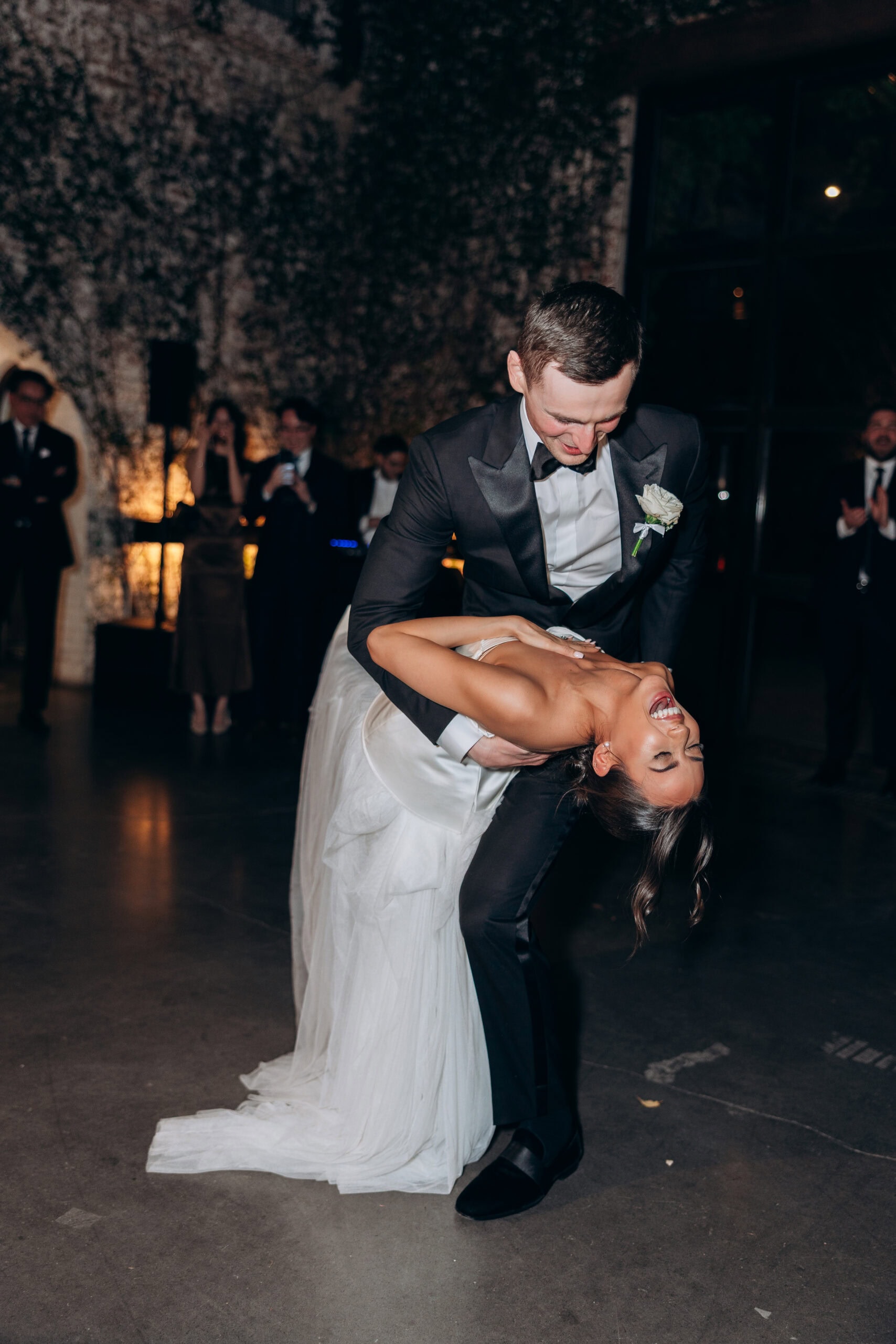 Bride in a draped white gown leans into a dramatic dance dip with the groom in a black tuxedo during their reception at The Foundry, while guests watch in the background.