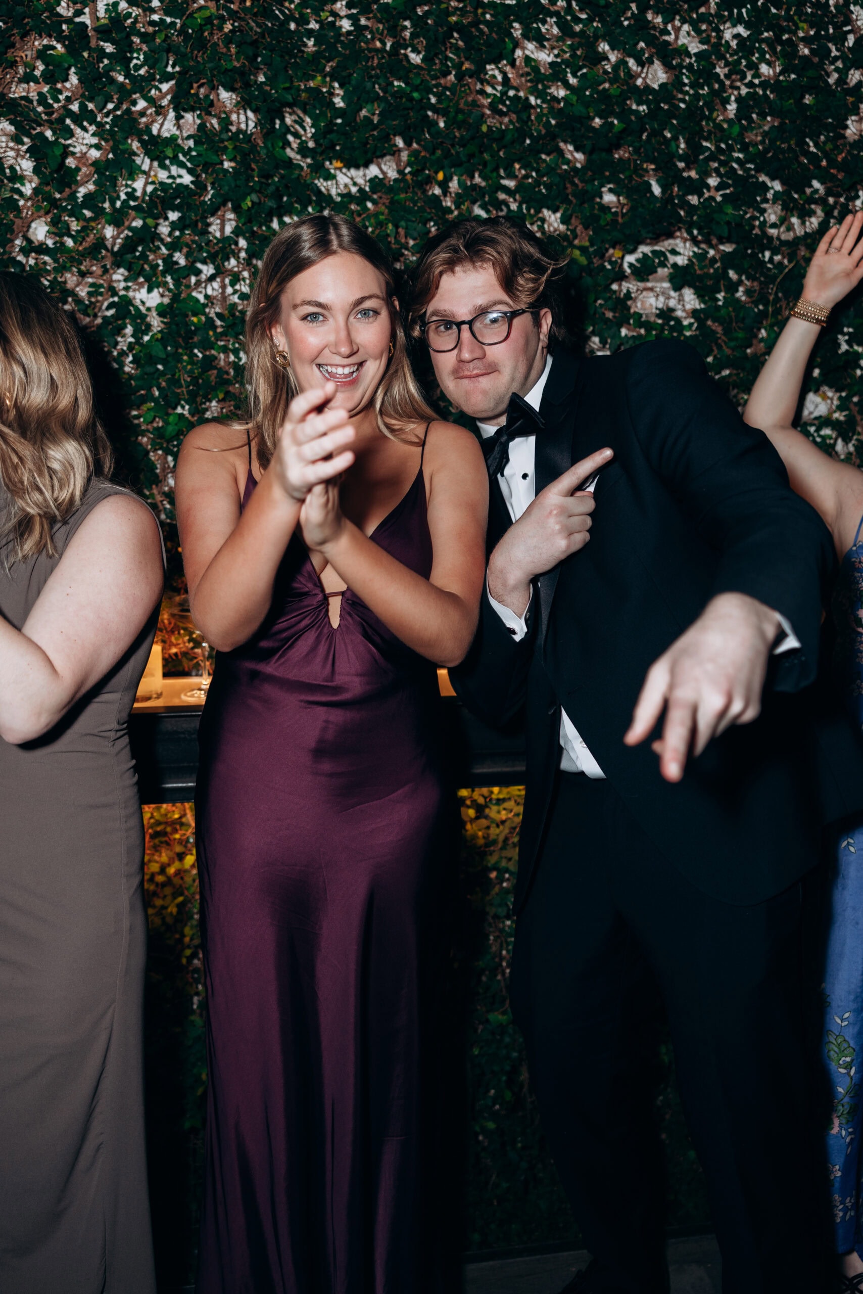 Two wedding guests pose playfully at an evening reception at The Foundry, standing before an ivy-covered wall in formal attire.