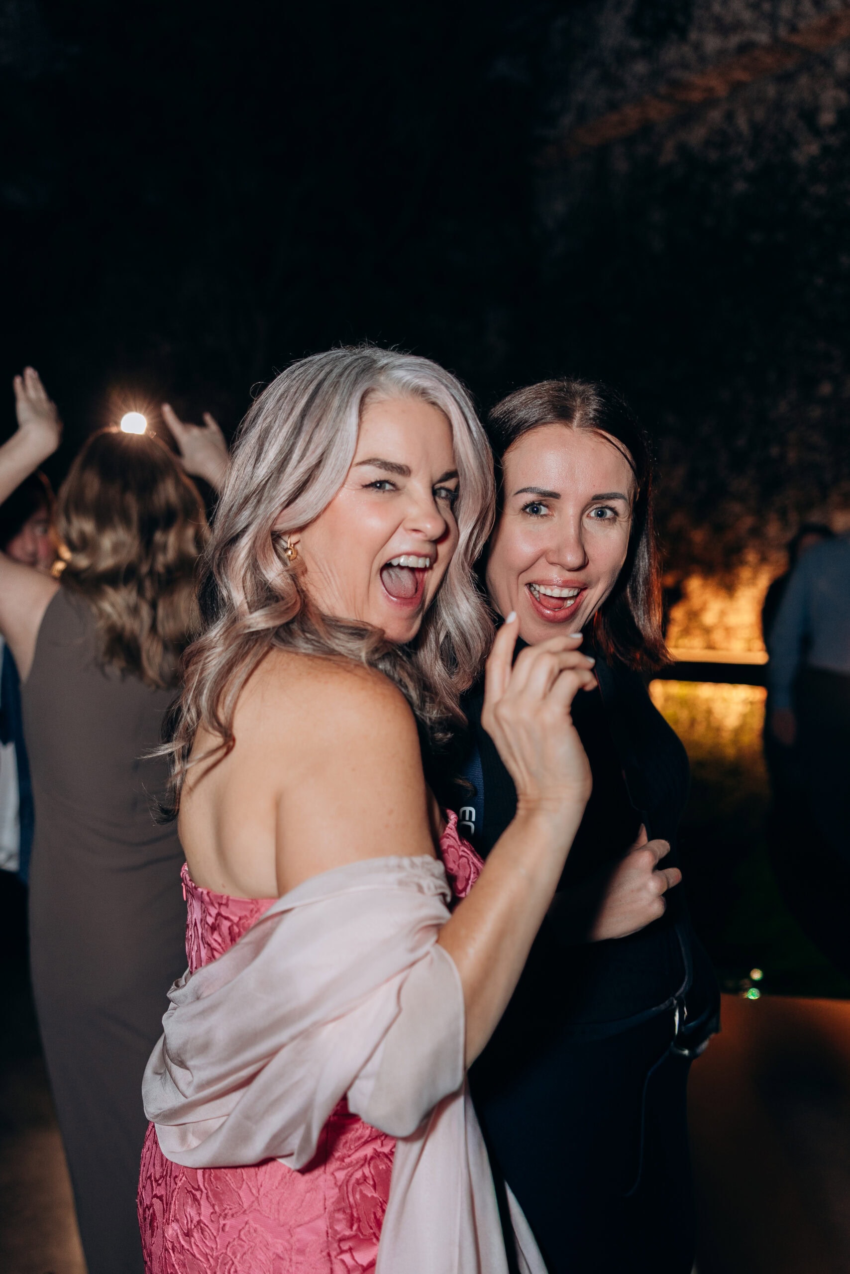 Two wedding guests smile and gesture toward the camera during an evening reception at The Foundry, with other guests dancing in the softly lit background.