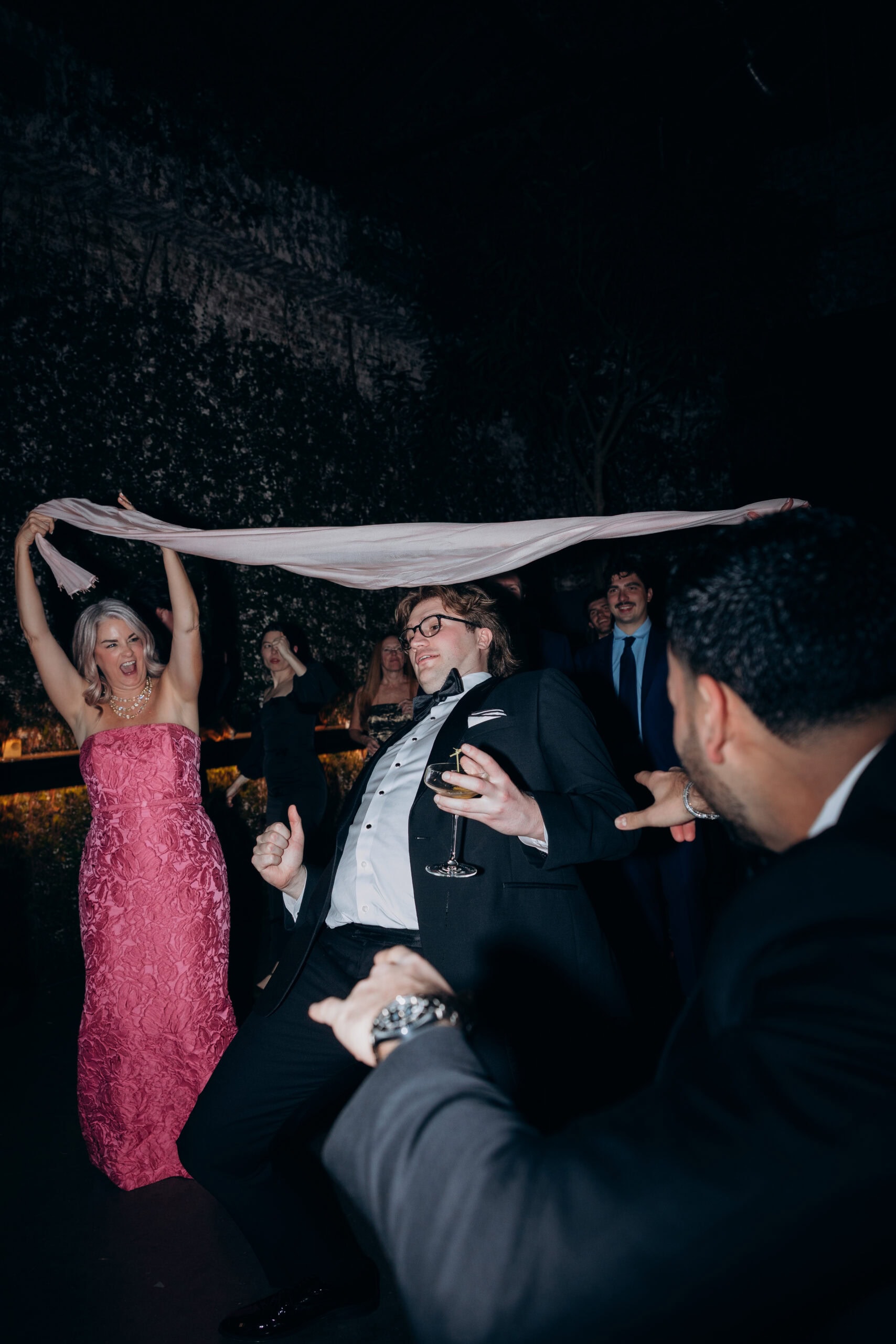 Nighttime wedding reception at The Foundry with a woman in a strapless pink gown holding a scarf overhead while a man in a tuxedo dances beneath it, surrounded by guests.