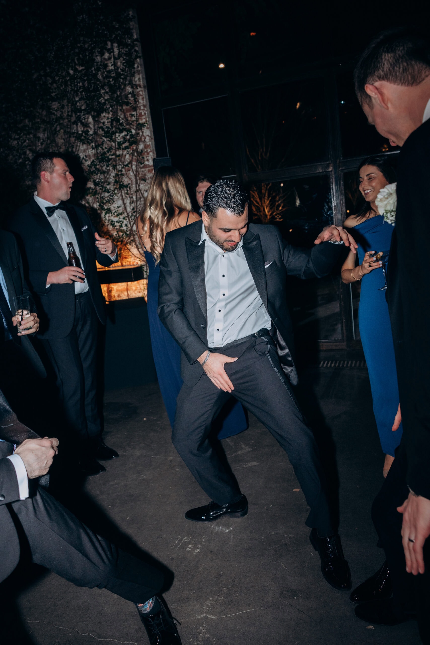 A man in a black tuxedo dances energetically at a wedding reception at The Foundry, surrounded by formally dressed guests holding drinks in a dark industrial space.