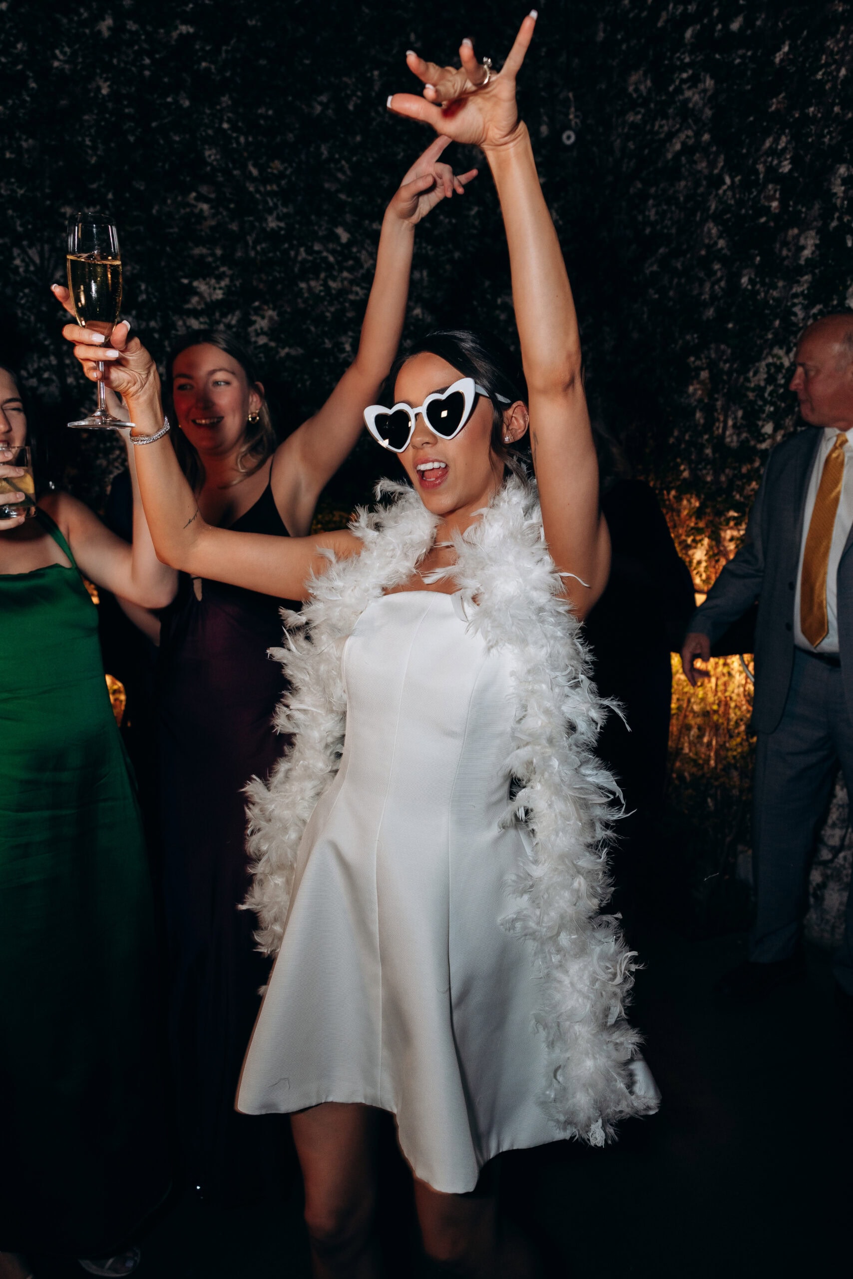 Bride dancing at The Foundry in a white mini dress, feather stole, and heart-shaped sunglasses, holding a champagne flute as guests celebrate around her.