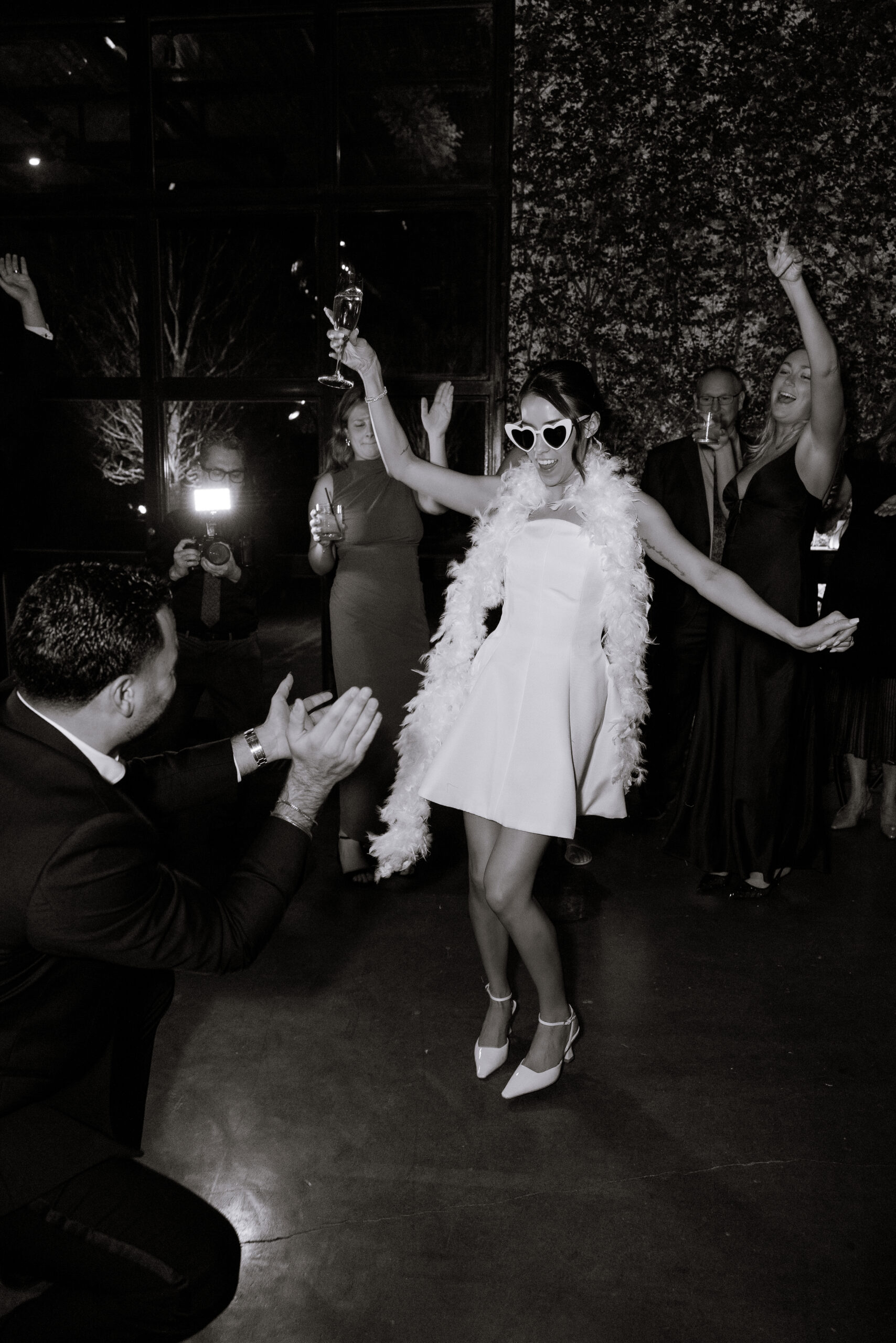Black-and-white wedding reception at The Foundry with a bride dancing in a short white dress, heart sunglasses, feather stole, and heels as guests clap around her.