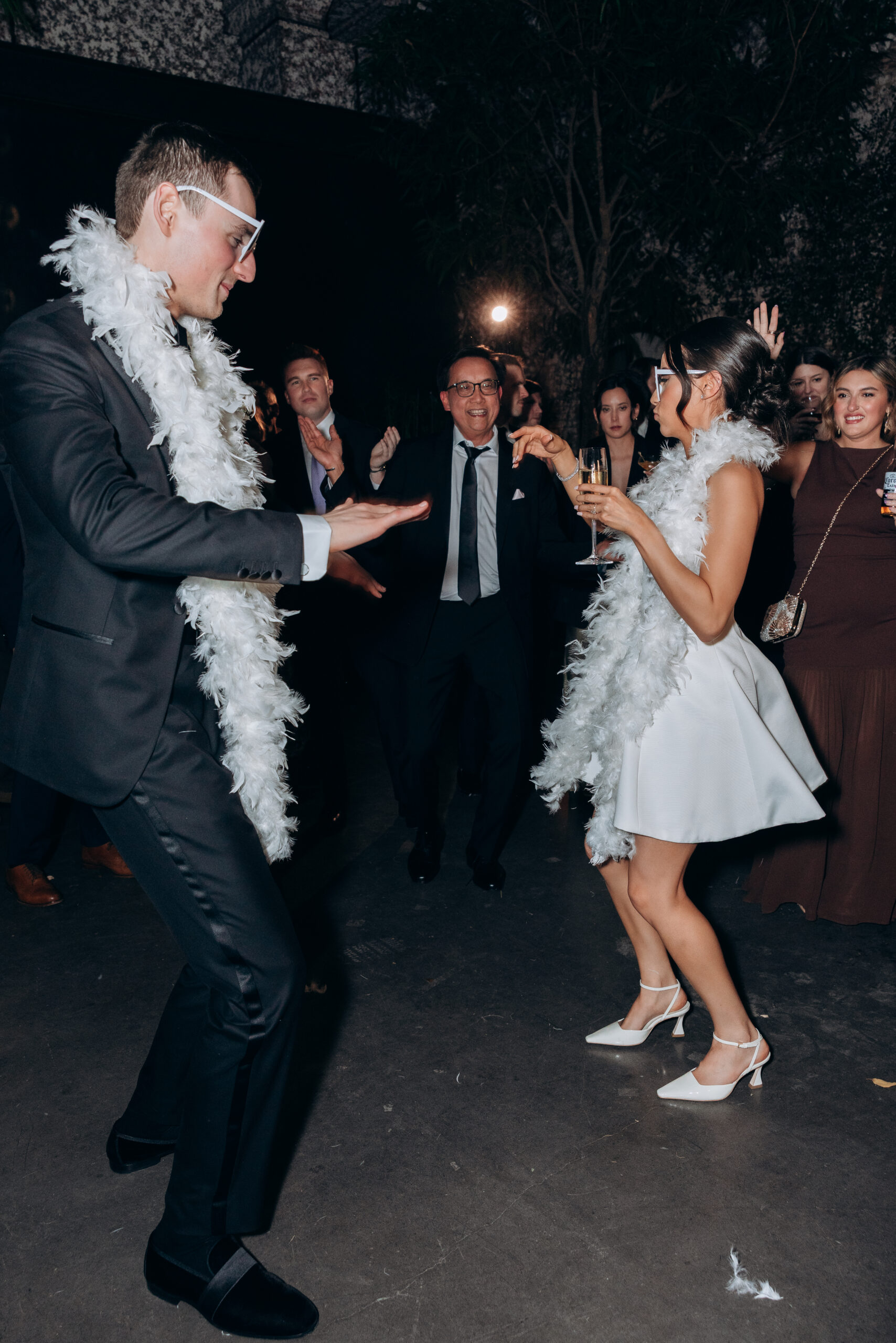 Bride and groom dance in white feather boas and party glasses at The Foundry, surrounded by guests during a lively late-night wedding reception.