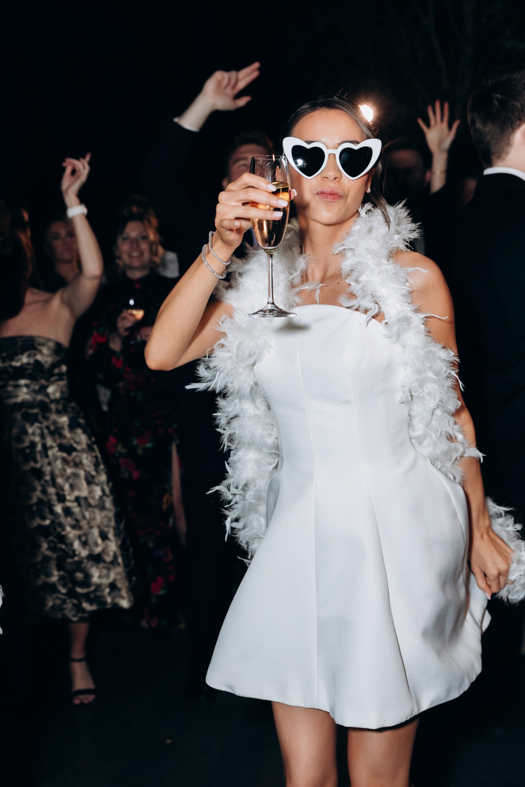 Bride in a white mini dress, feather stole, and heart-shaped sunglasses toasts with champagne while dancing at a wedding reception at The Foundry.