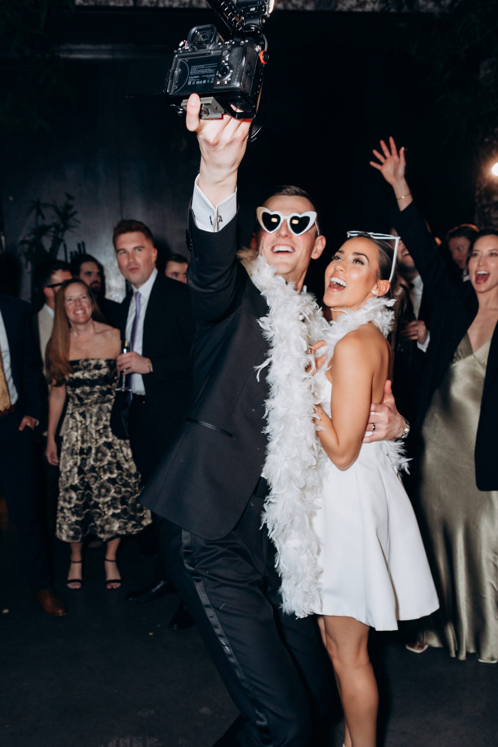 At The Foundry, a couple in formal wedding attire pose on the dance floor as one lifts a camera overhead; both wear novelty sunglasses and feather boas while guests cheer behind them.