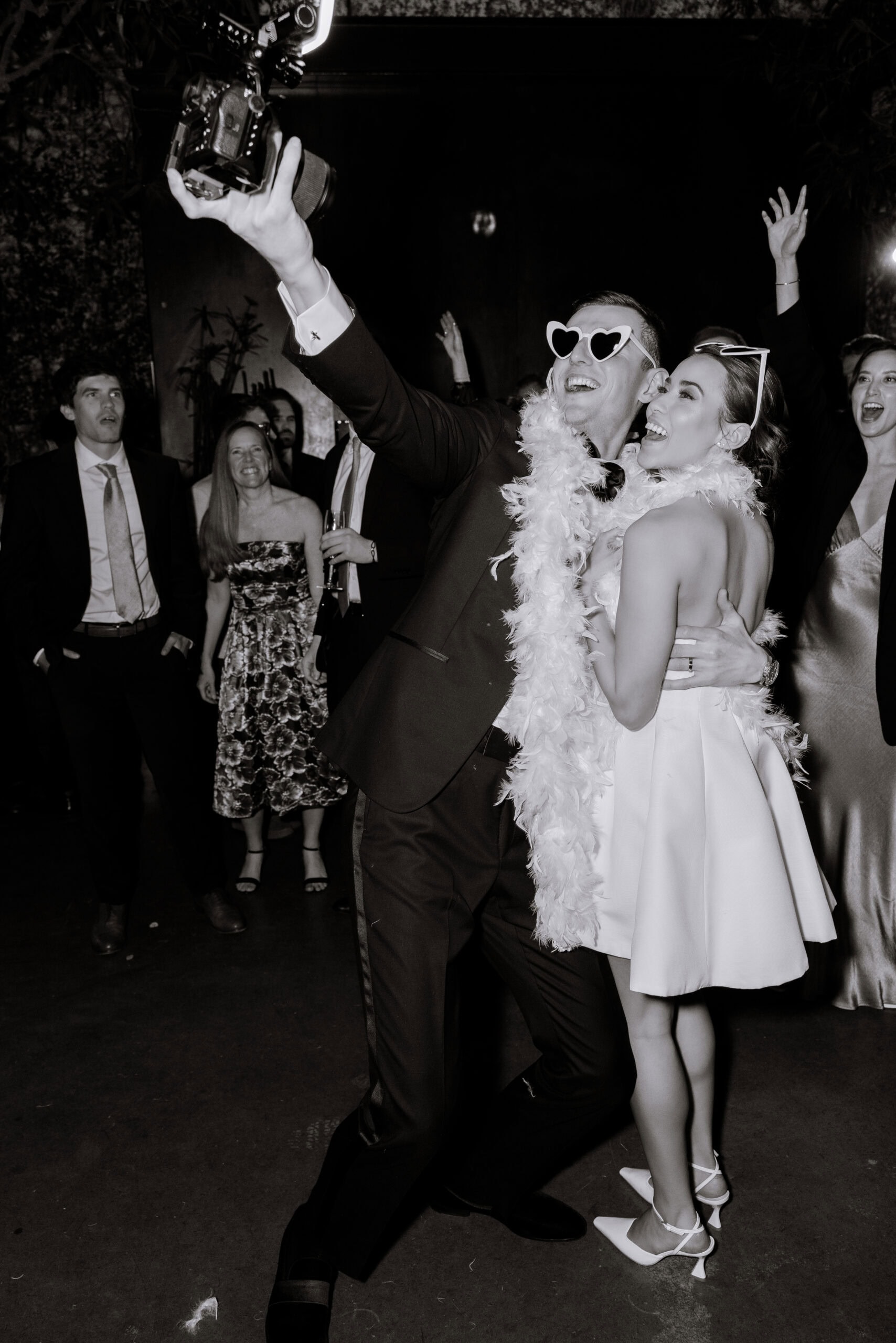 Black-and-white wedding reception photo at The Foundry showing a couple posing for a selfie with a camera, wearing heart-shaped sunglasses and feather boas as guests cheer behind them.