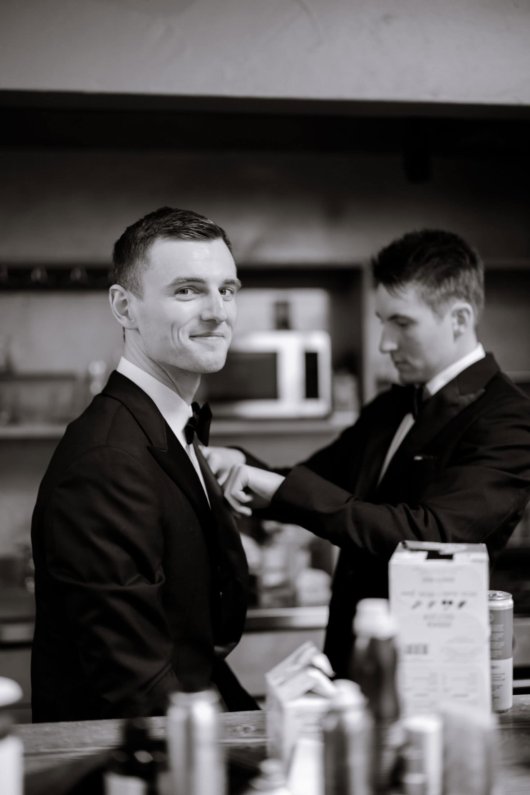 Black-and-white image of two men in tuxedos during wedding preparations at The Foundry, with one smiling at the camera while the other adjusts his jacket.