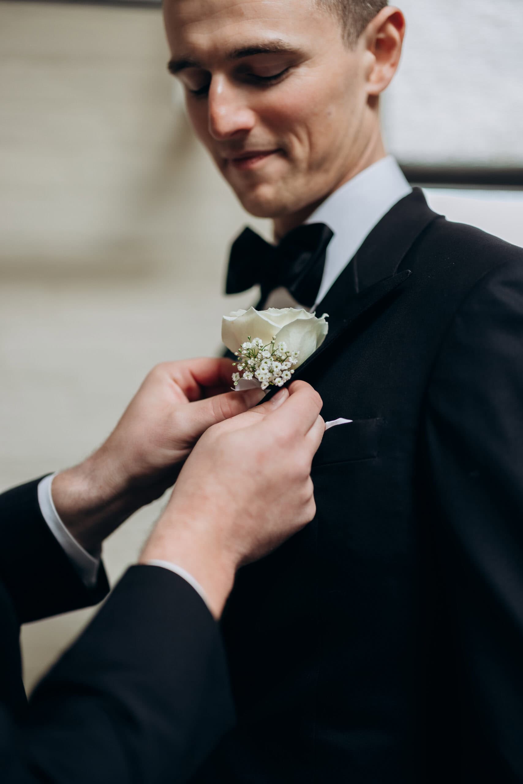Close-up of a groom in a black tuxedo as another person pins a white rose and baby's breath boutonniere to his lapel at The Foundry.