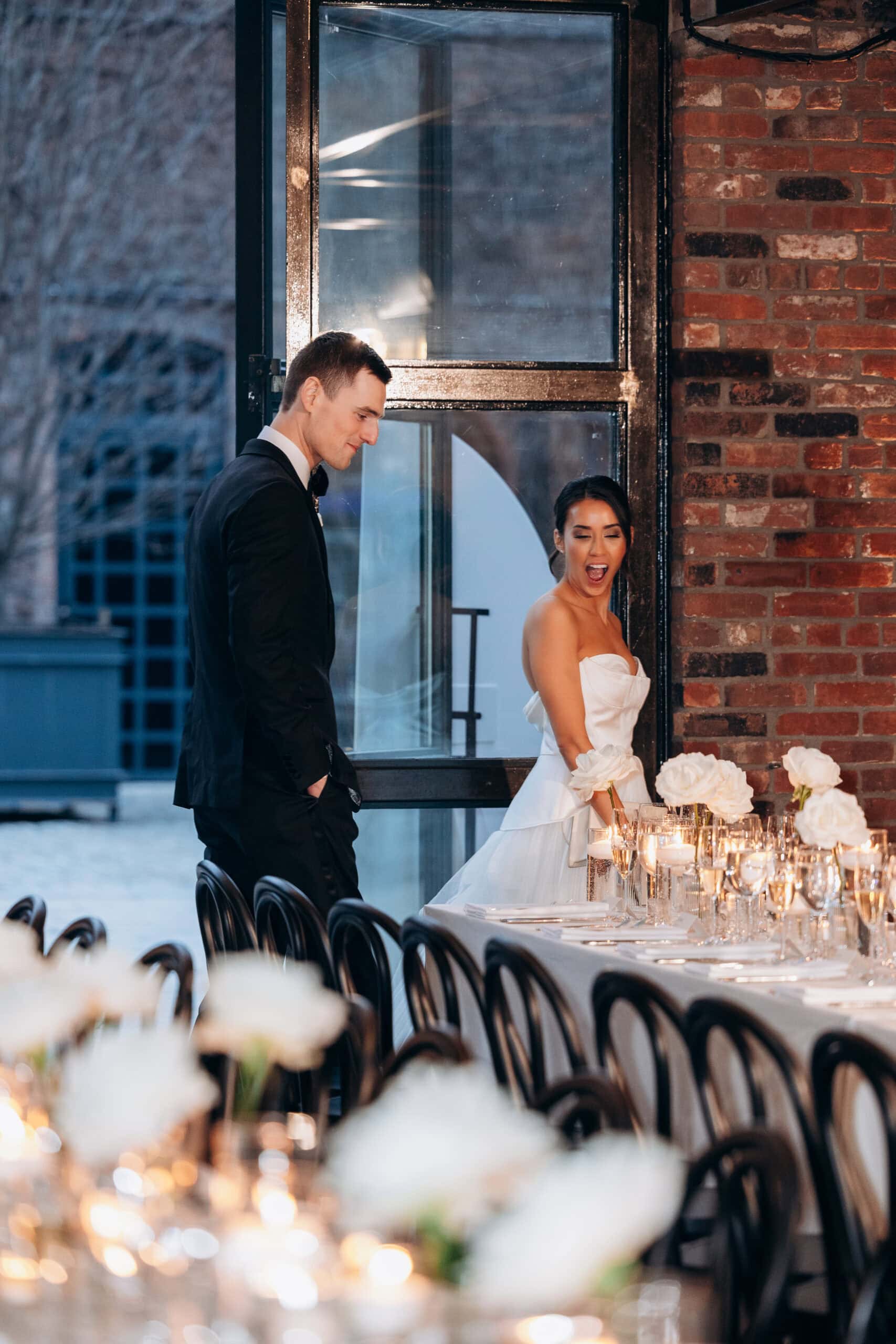 Bride and groom pause beside a candlelit reception table with white roses inside The Foundry, framed by brick walls and tall industrial windows at dusk.