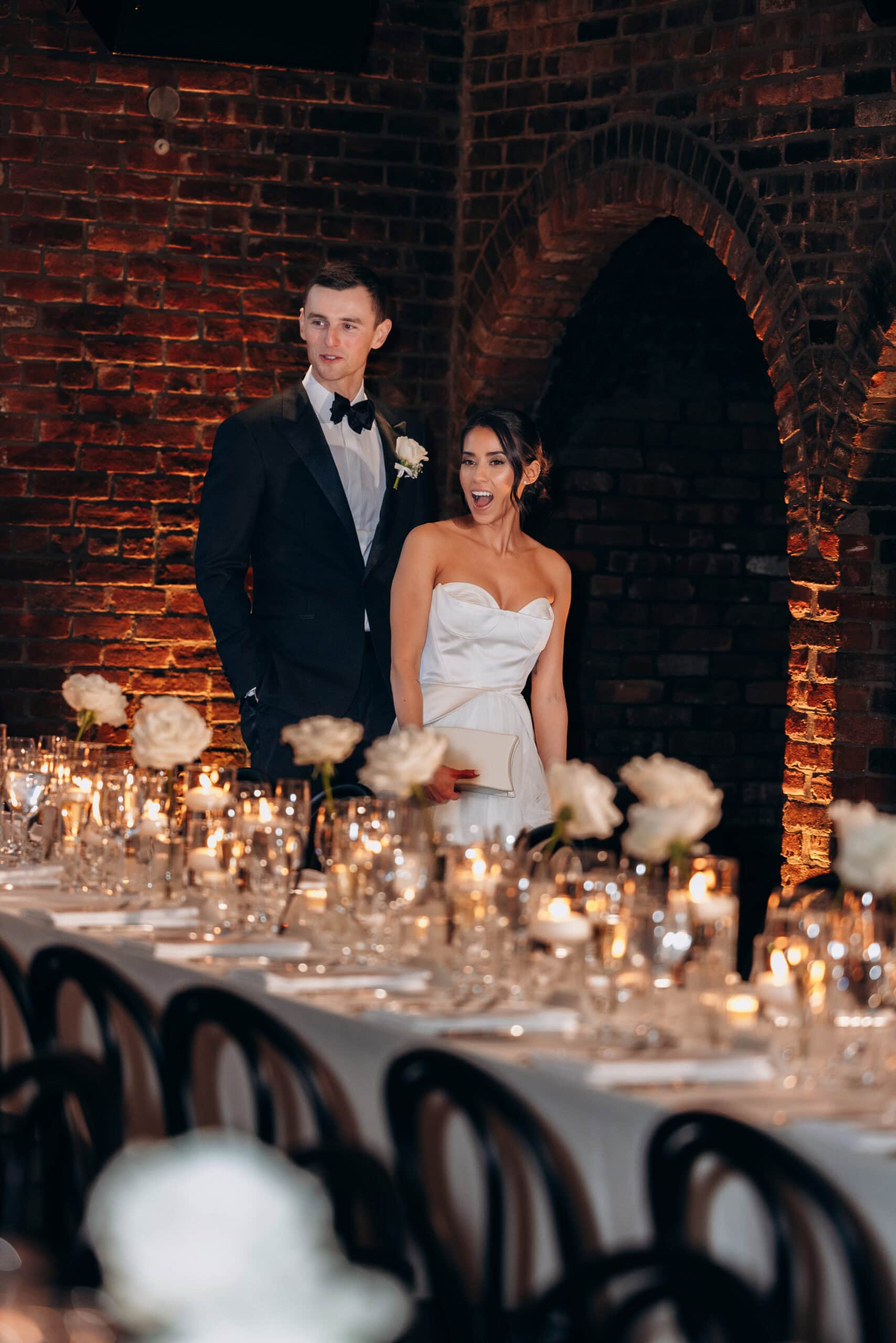 Bride and groom enter a candlelit reception at The Foundry, framed by exposed brick arches, white rose centerpieces, and a long table set with glassware.