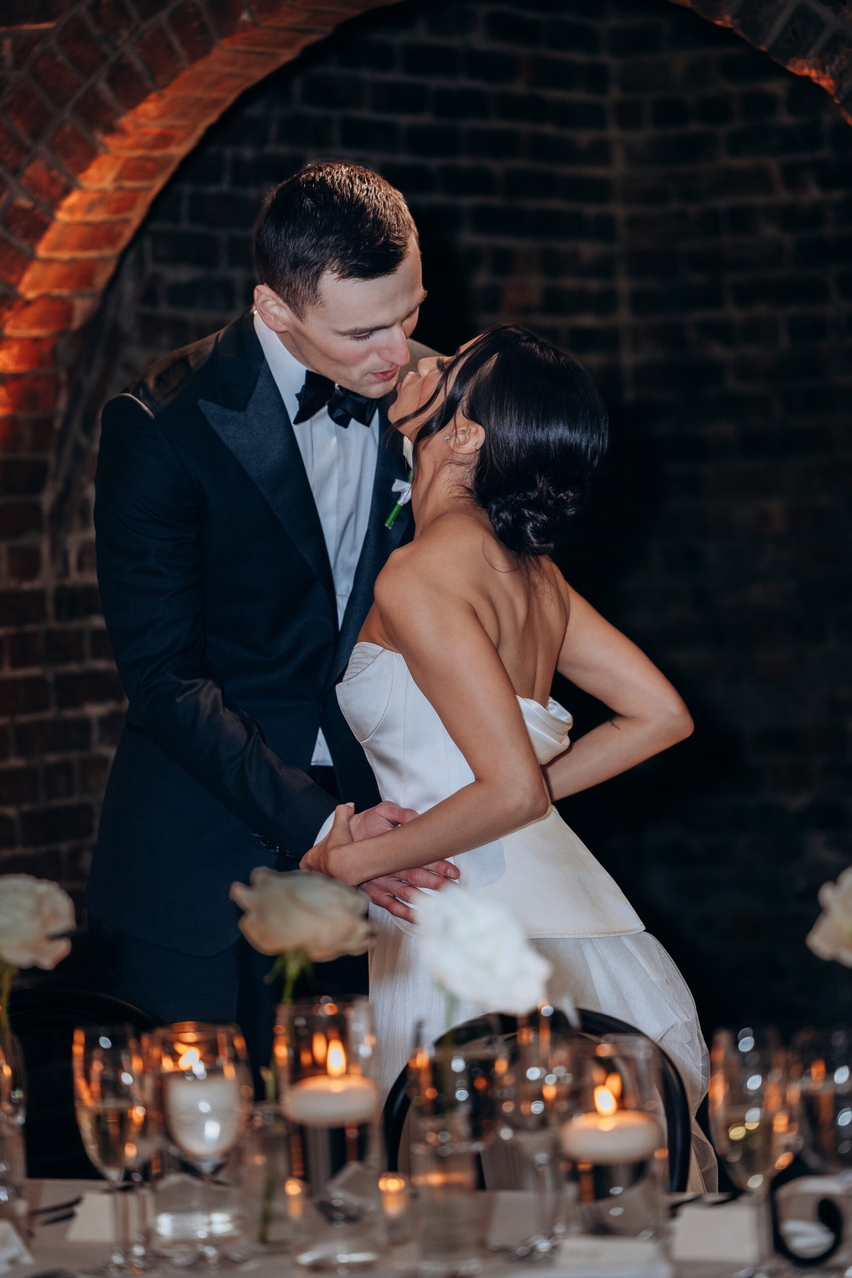 Bride and groom share a kiss at their reception beneath a brick arch at The Foundry, surrounded by candlelit tables, glassware, and white florals.