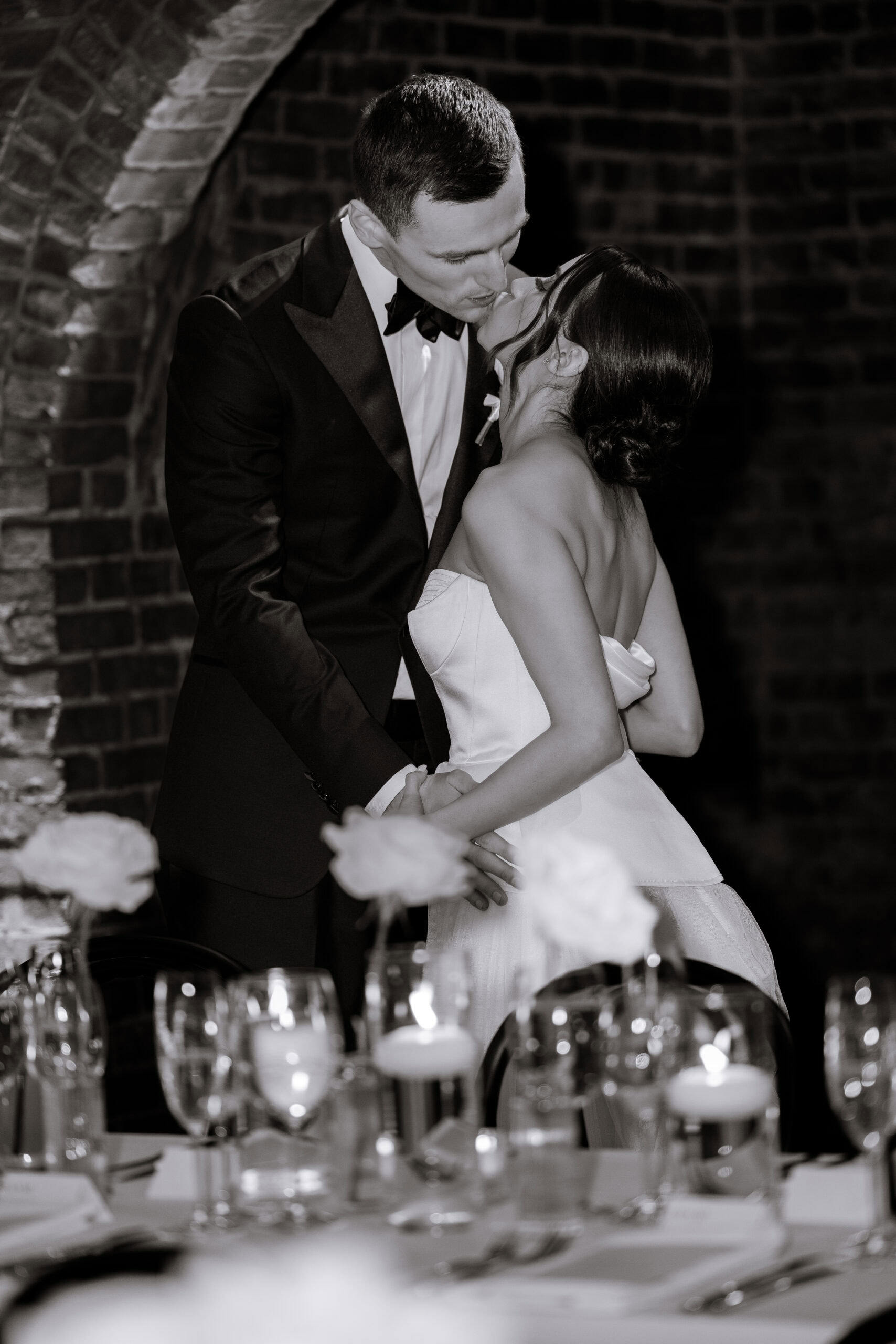 Black-and-white wedding portrait of a couple kissing at their reception at The Foundry, framed by brick arches, candlelit tables, glassware, and white flowers.
