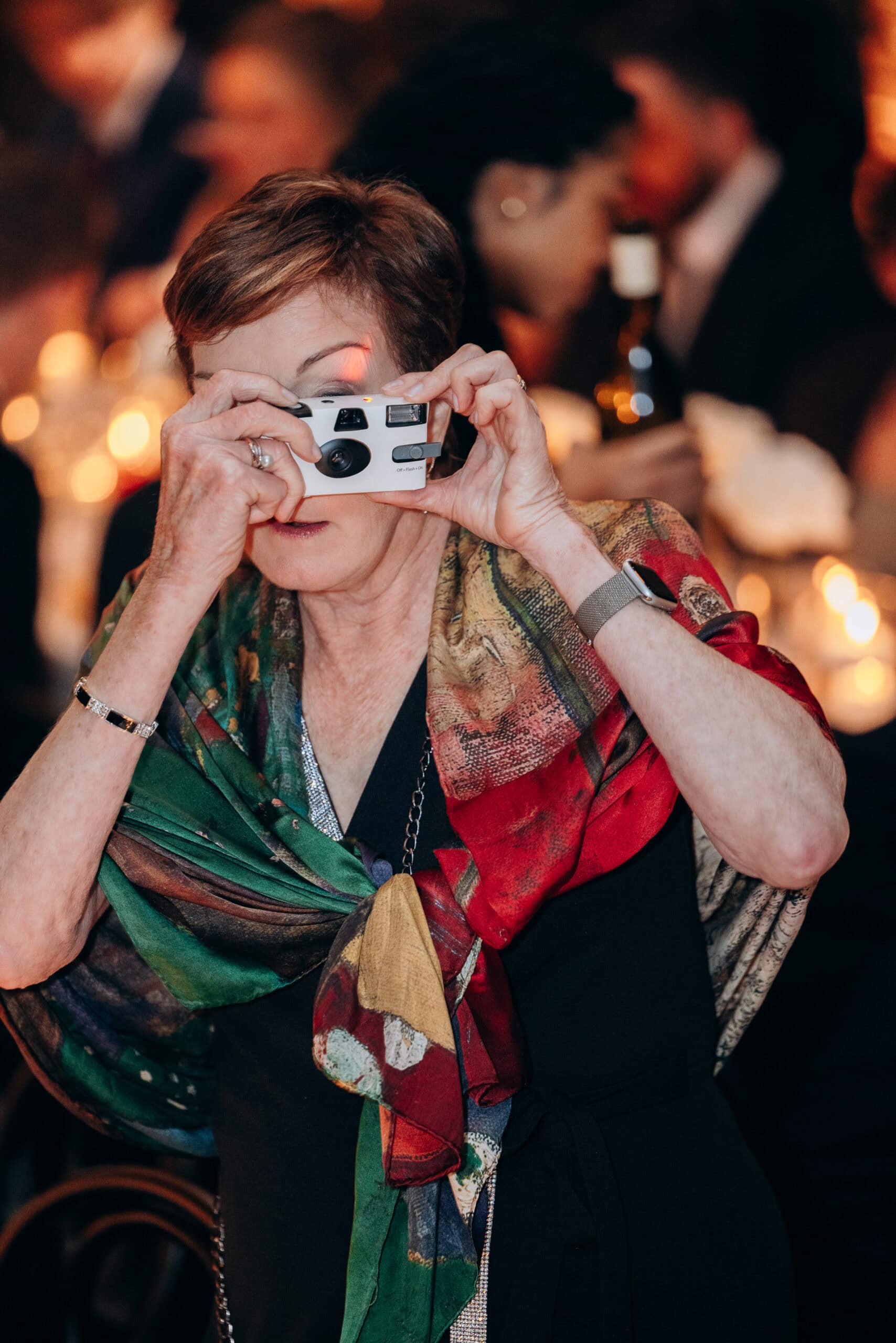 At The Foundry, an older wedding guest in a black dress and multicolored silk wrap holds a disposable camera to take a photo during a candlelit reception.