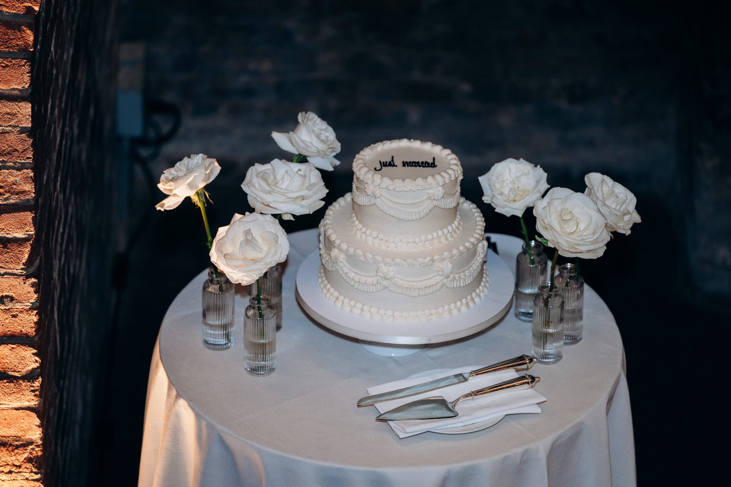 Ivory two-tier wedding cake with piped garlands and “Just married” icing on a round table at The Foundry, surrounded by white roses in glass bud vases.