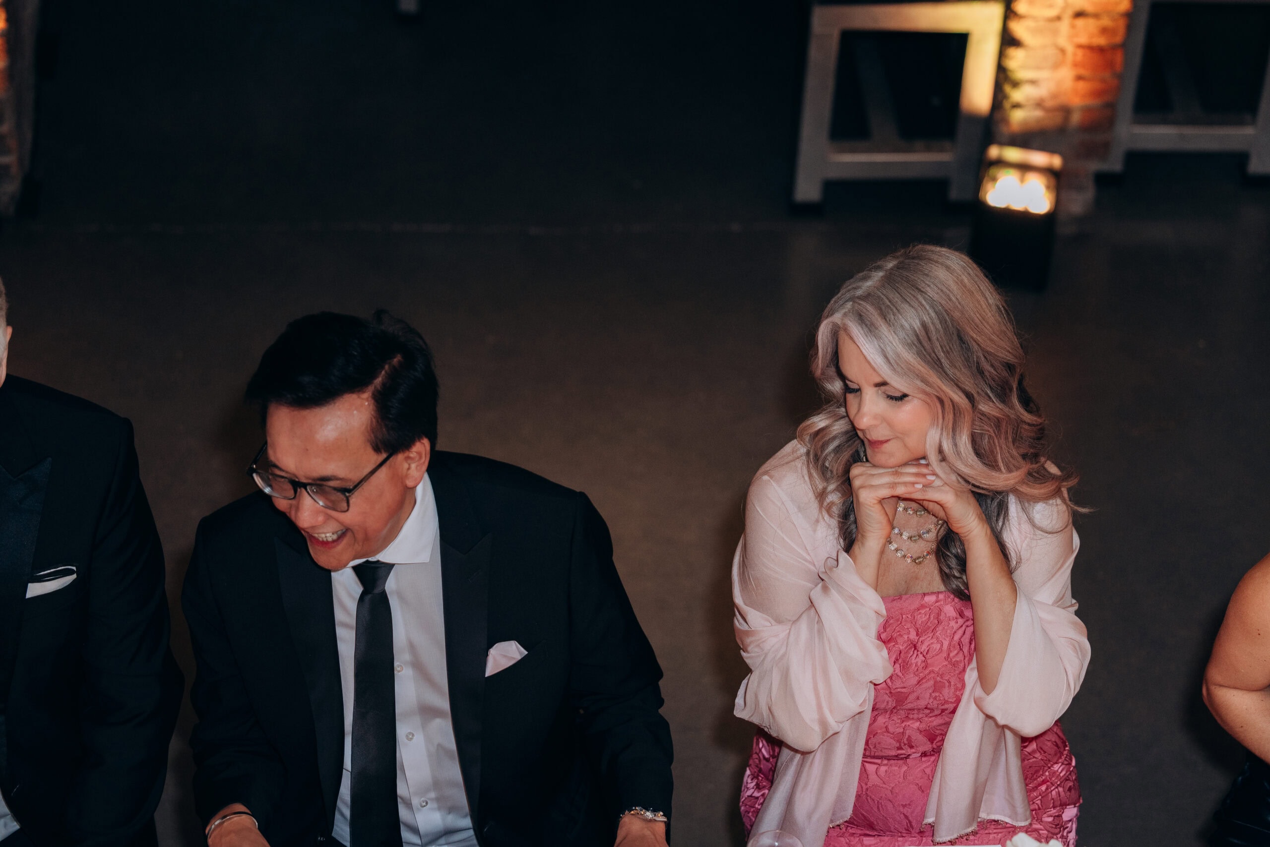 Two formally dressed wedding guests seated indoors at The Foundry during a candlelit reception, with a man in a black suit and a woman in a pink textured dress and shawl.