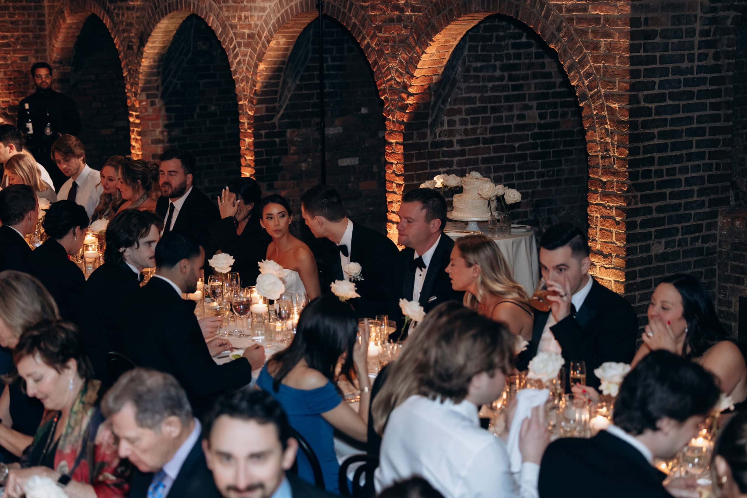 Wedding reception at The Foundry with guests seated at candlelit tables beneath brick arches, with white roses, glassware, and a tiered cake in the background.