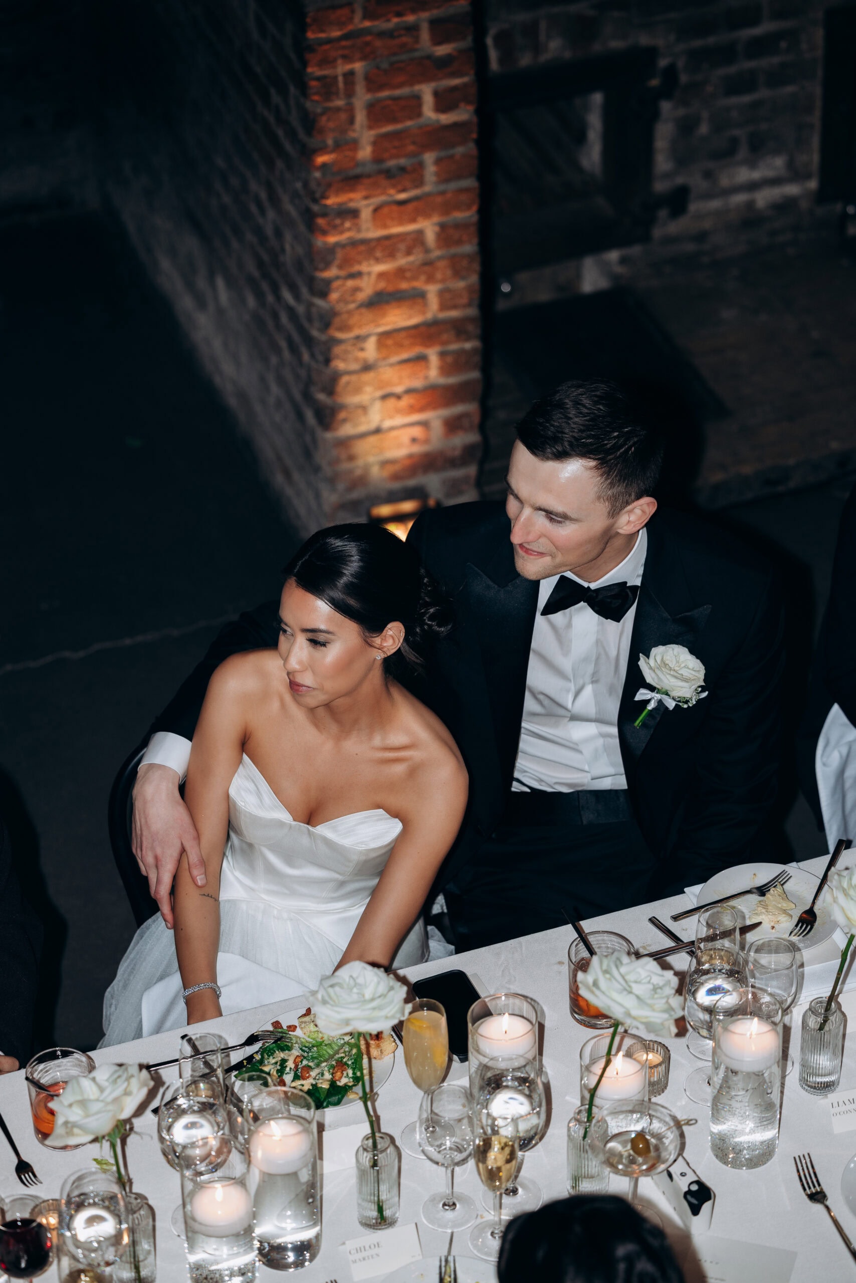 Overhead view of a wedding couple seated closely at a candlelit reception table at The Foundry, with brick walls, white roses, crystal glassware, and plated dinner.