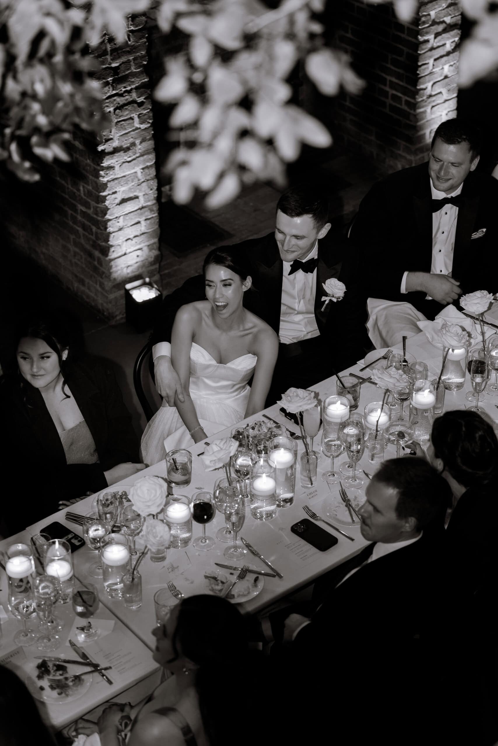 Black-and-white overhead photo of a bride and groom seated at a candlelit reception table with guests, white roses, and brick walls at The Foundry.