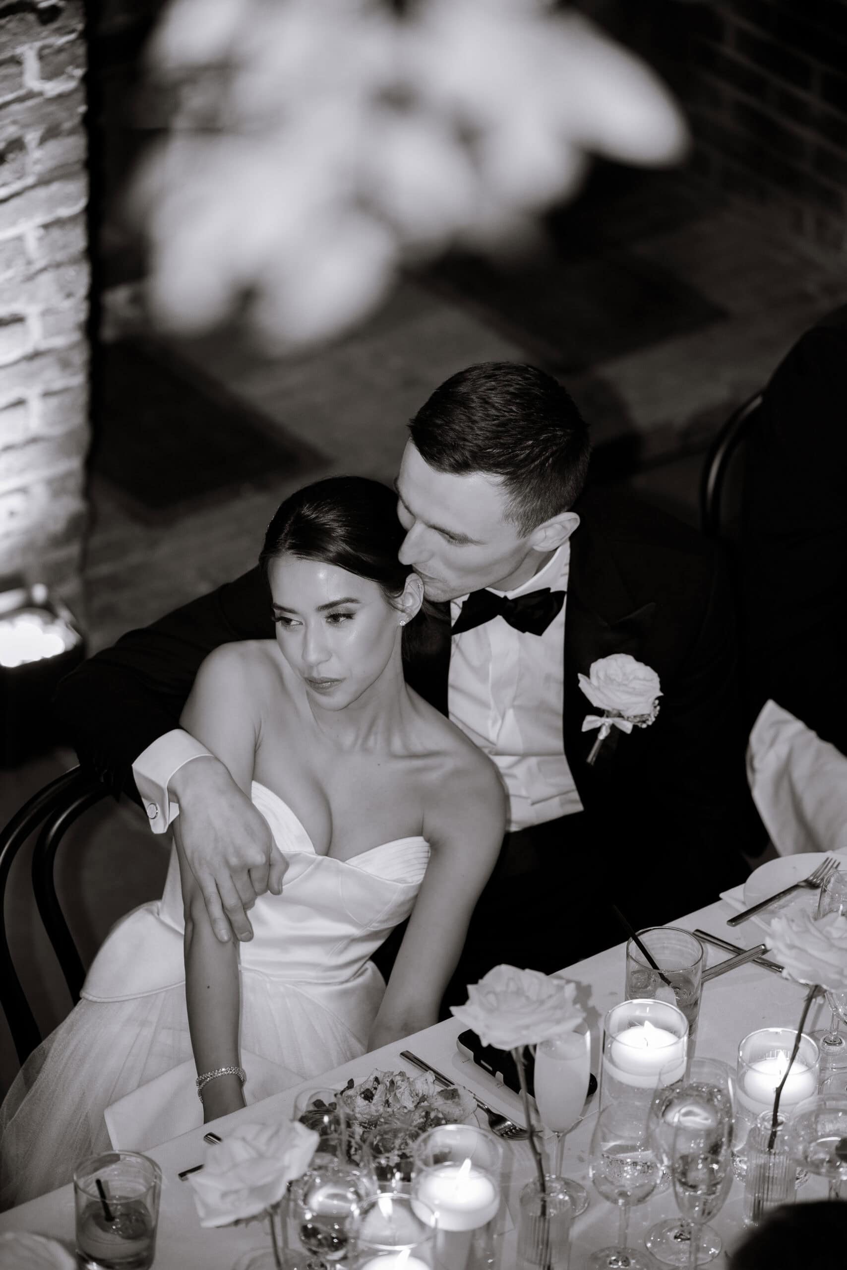 Black-and-white wedding reception portrait at The Foundry of a seated bride in a strapless satin gown and a groom in a tuxedo embracing her at a candlelit table.